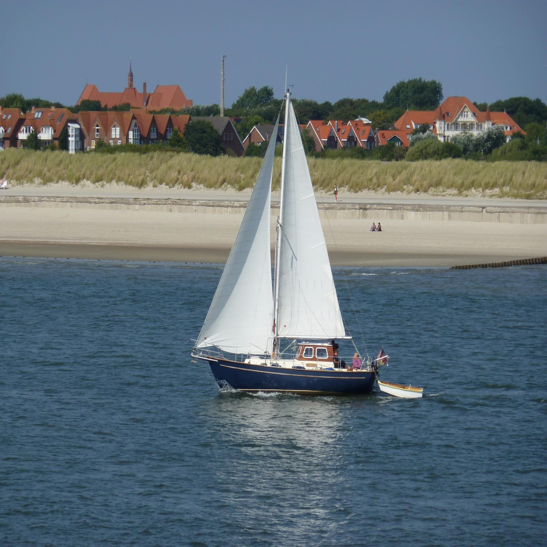 Ein kleines Segelboot schippert vor dem Strand, Dünen und Häuser sind im Hintergrund zu sehen. 
