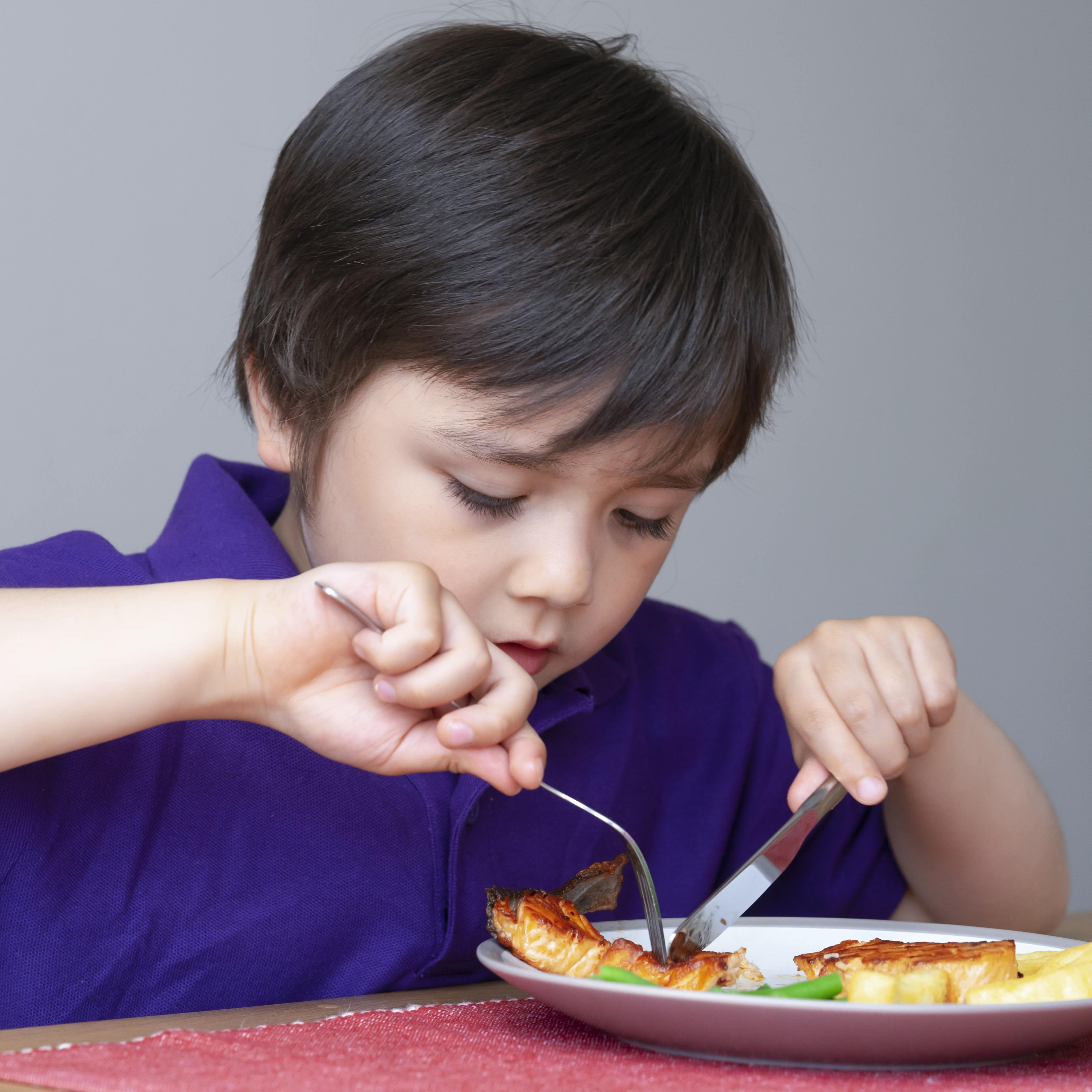 Ein kleiner Junge im lila Polohemd sitzt am Tisch und schneidet sein Essen: Gegrillter Frosch, Pommes und Gemüse.