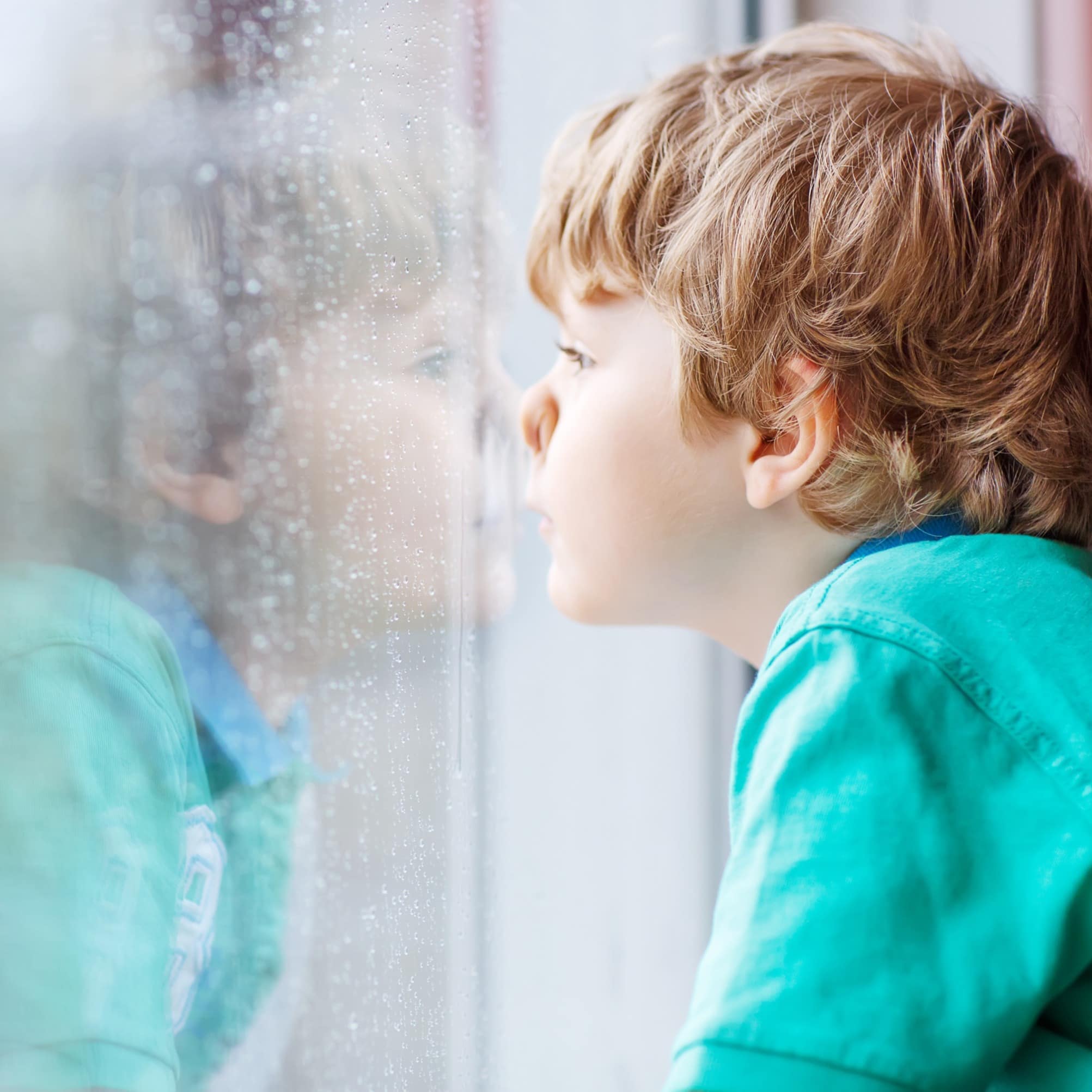 Kleiner Junge in grünem T-Shirt drückt die Nase an ein Fenster und schaut in den Regen.