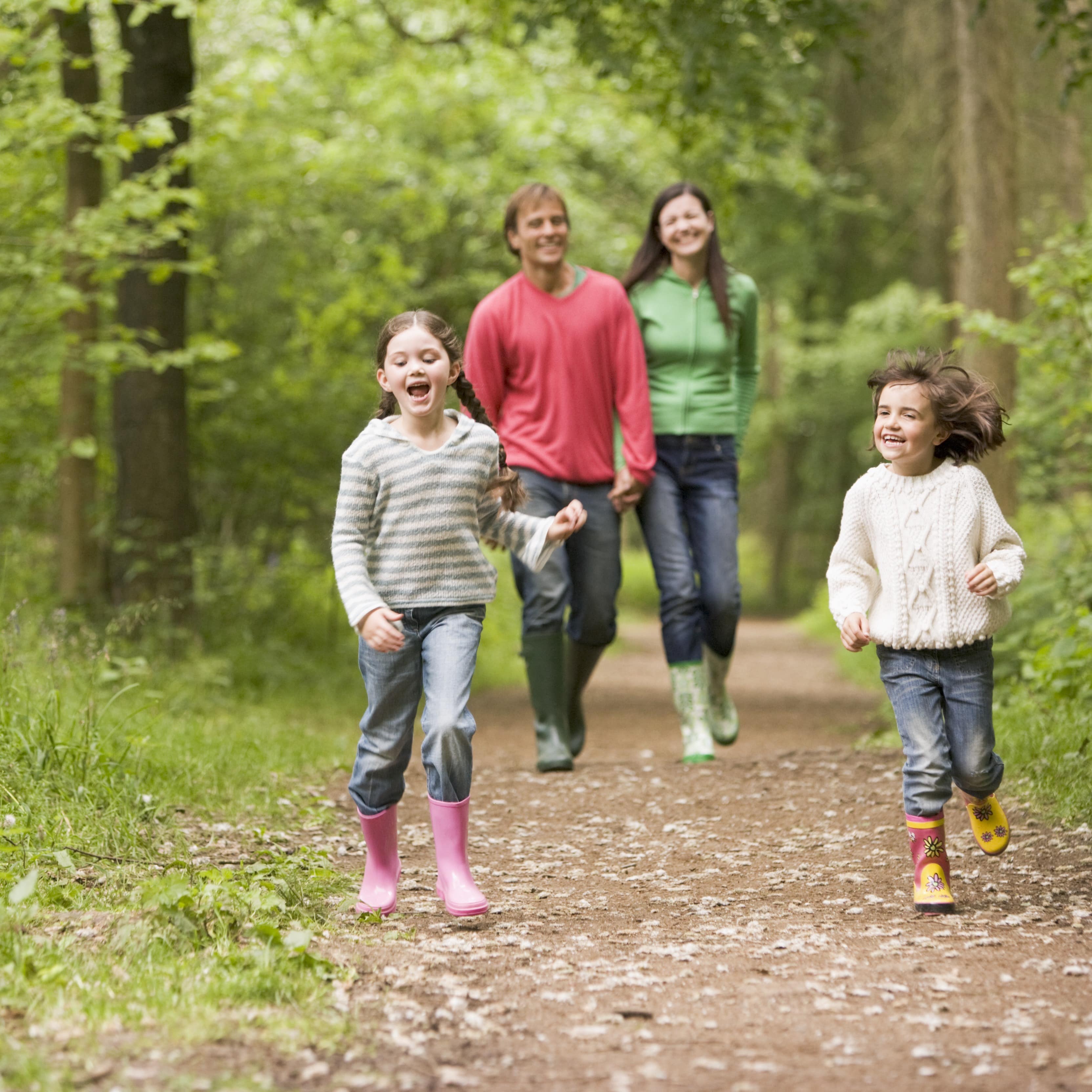 2 Kinder laufen einen Waldweg entlang, im Hintergrund die Eltern. 