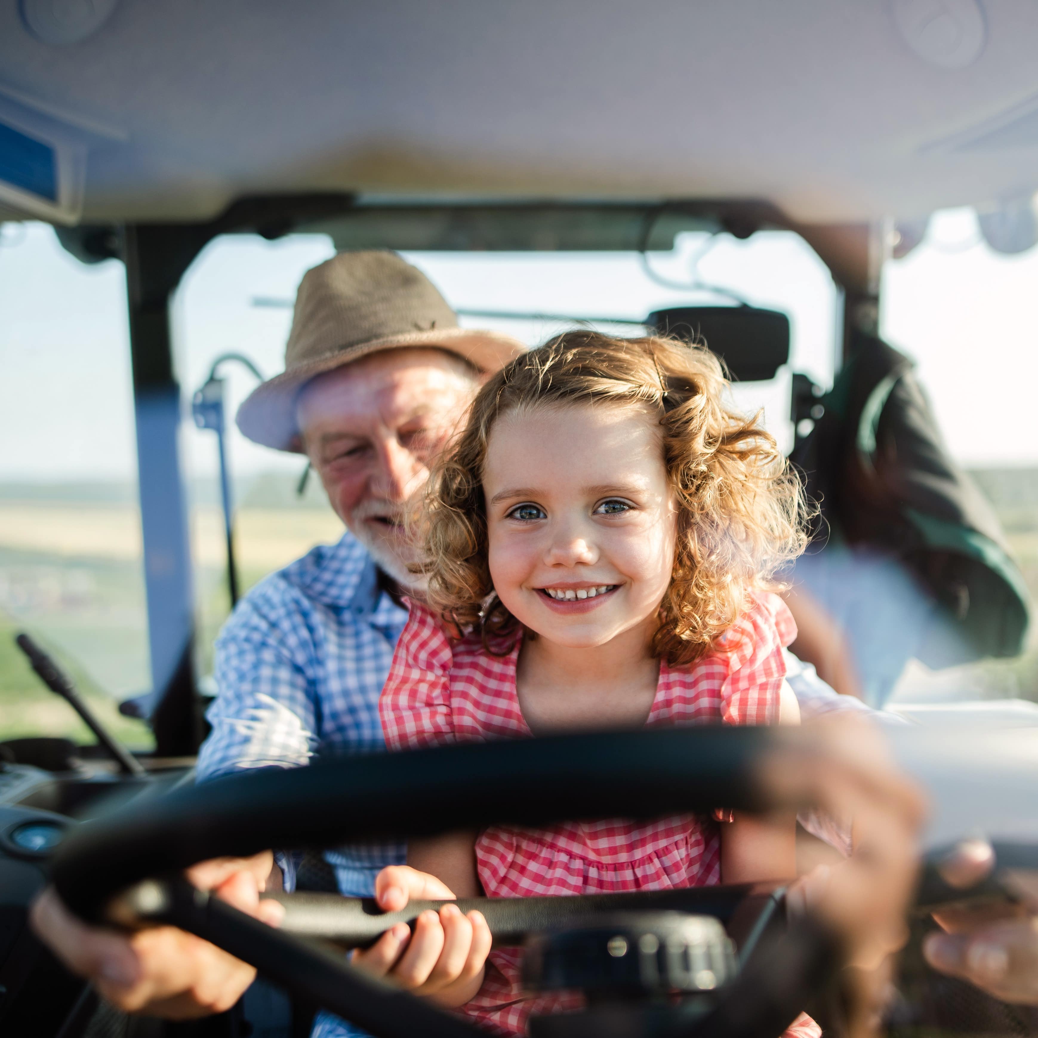 Ein kleines Mädchen sitzt bei einem Bauern auf dem Schoß am Steuer eines Traktors. Beide tragen karierte Sommerkleidung.