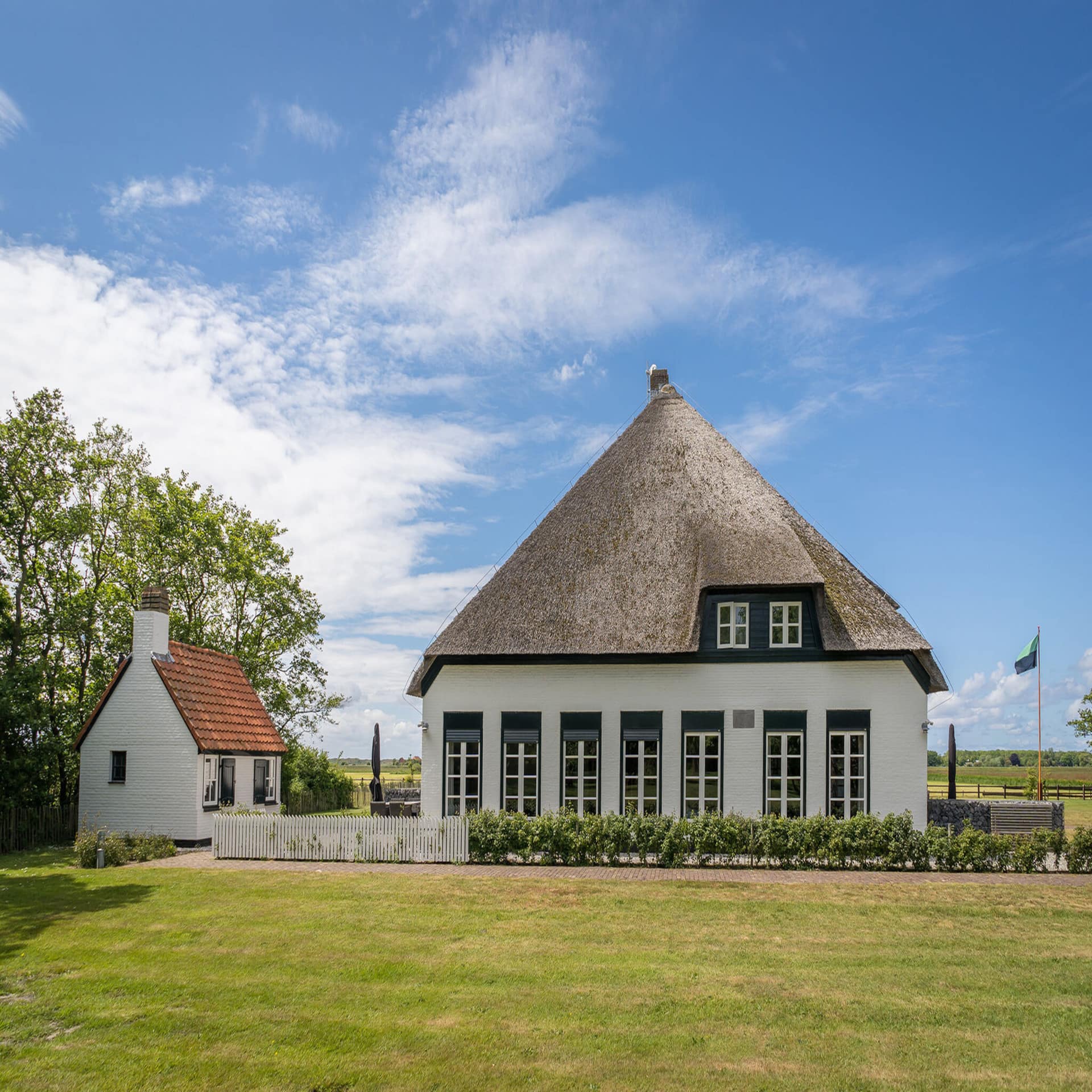Ein weißes Reetdachhaus mit Garten, dahinter grünes Land. Der Himmel ist blau, die Sonne scheint.