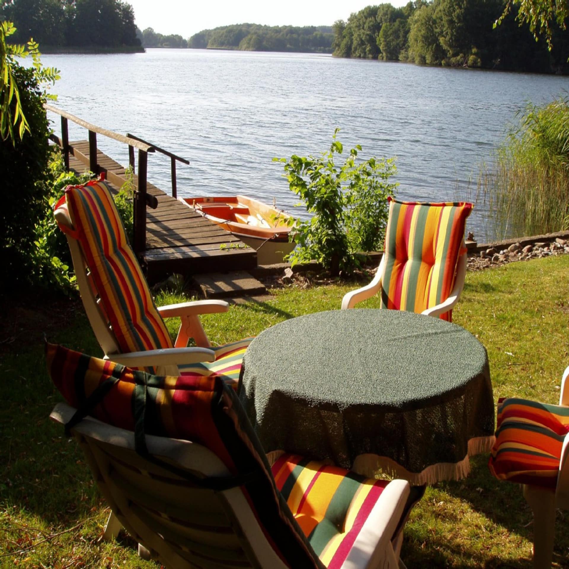 Gartentisch und 4 Stühle in einem Garten direkt am Wasser. Ein Bootssteg und ein Ruderboot sind zu sehen. Die Sonne scheint.
