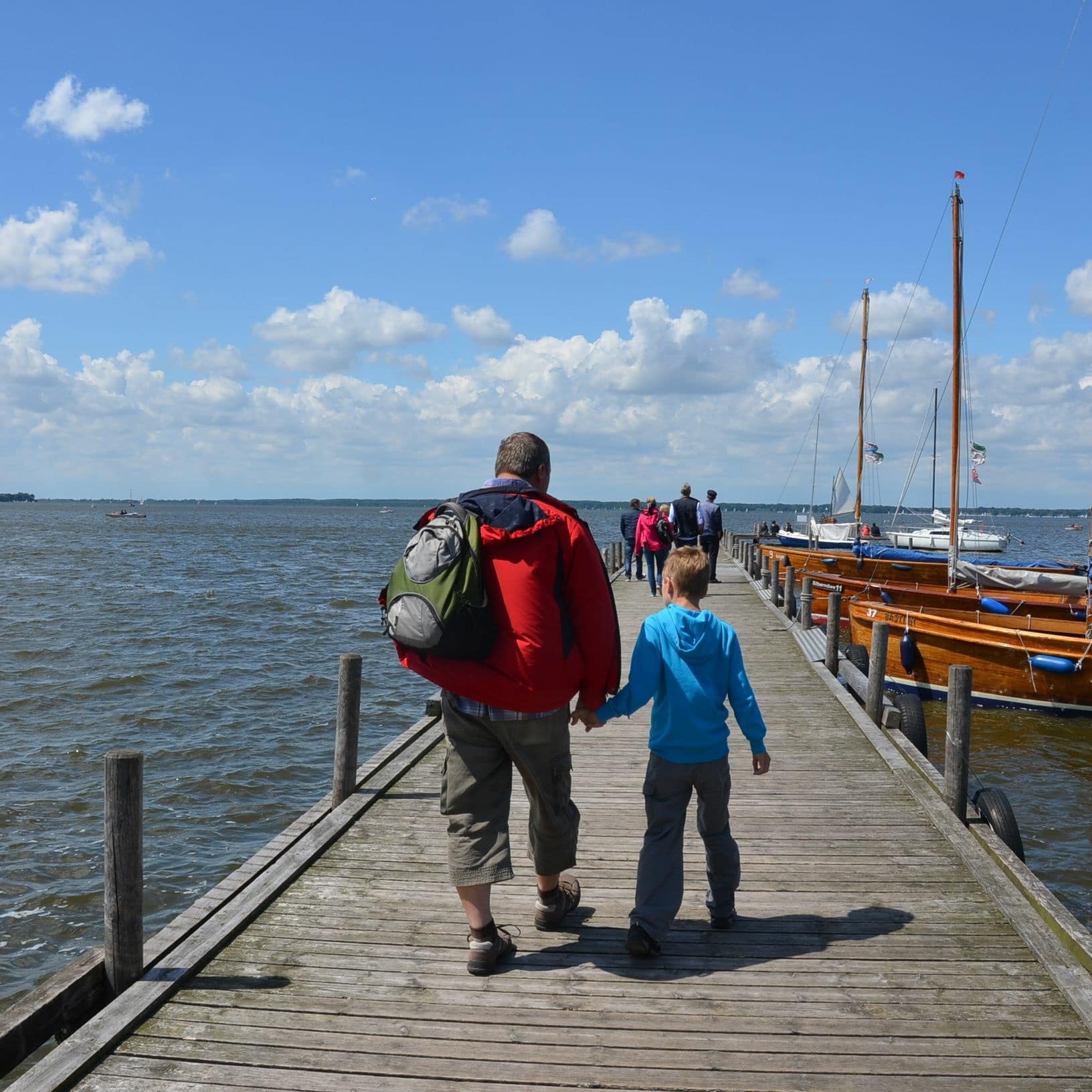 Rückenansicht: Vater mit einem Jungen an der Hand auf einem Bootssteg am Steinhuder Meer, rechts einige Segelboote.