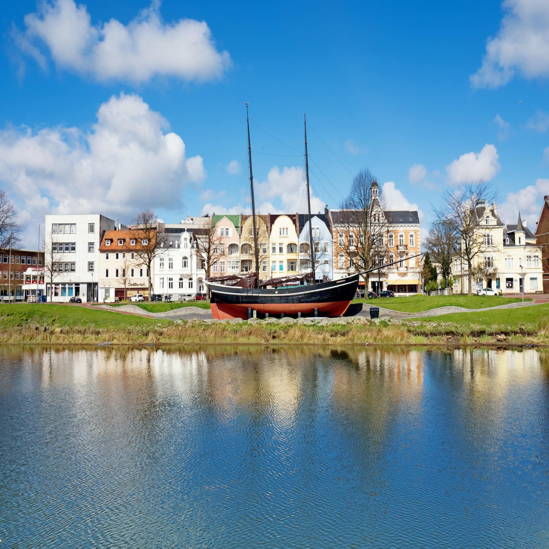 Blick von Wasser auf einen Park mit Segelboot und Gebäuden im Hintergrund.