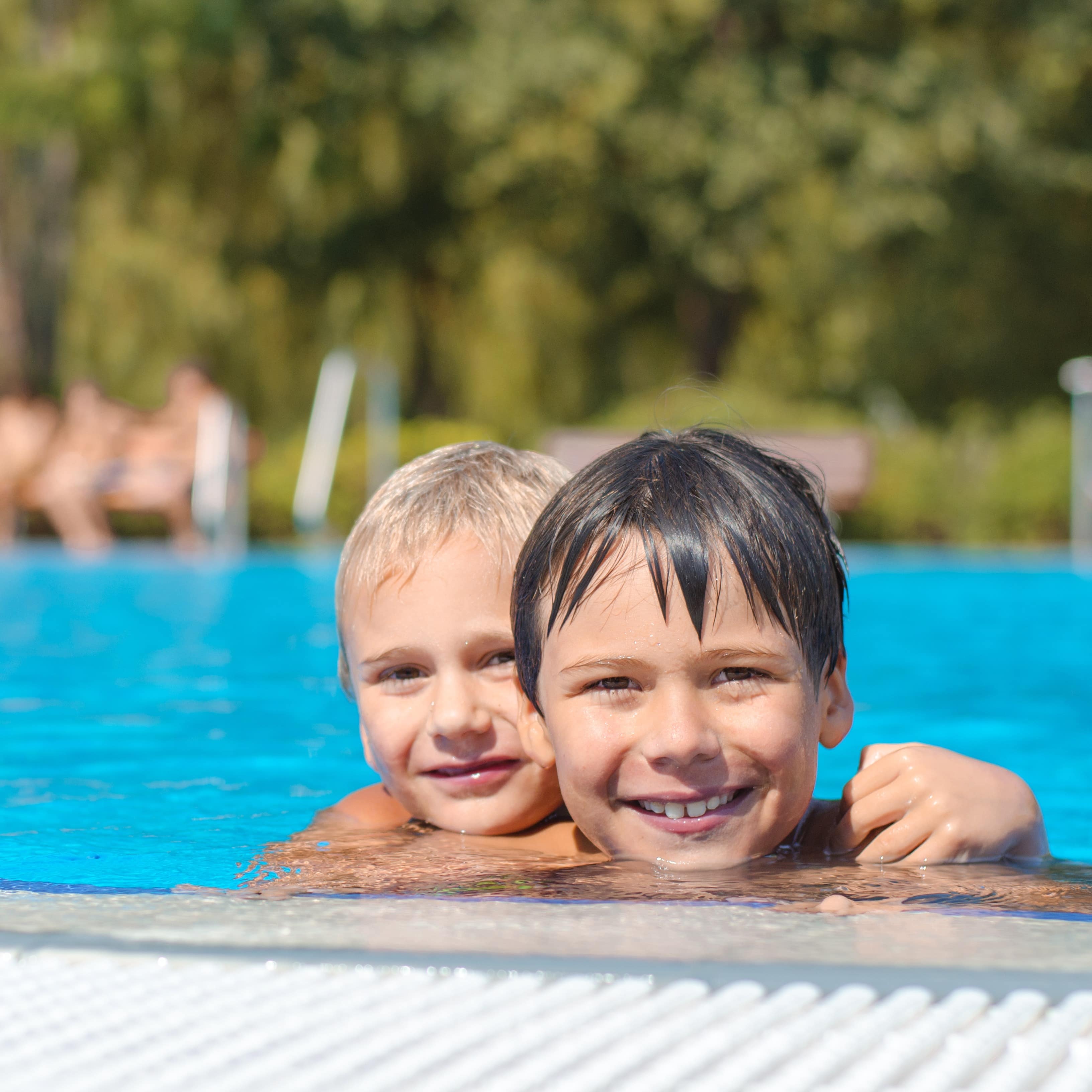 2 Jungen im Freibad schauen aus dem Wasser eines Freibads und lachen in die Kamera.