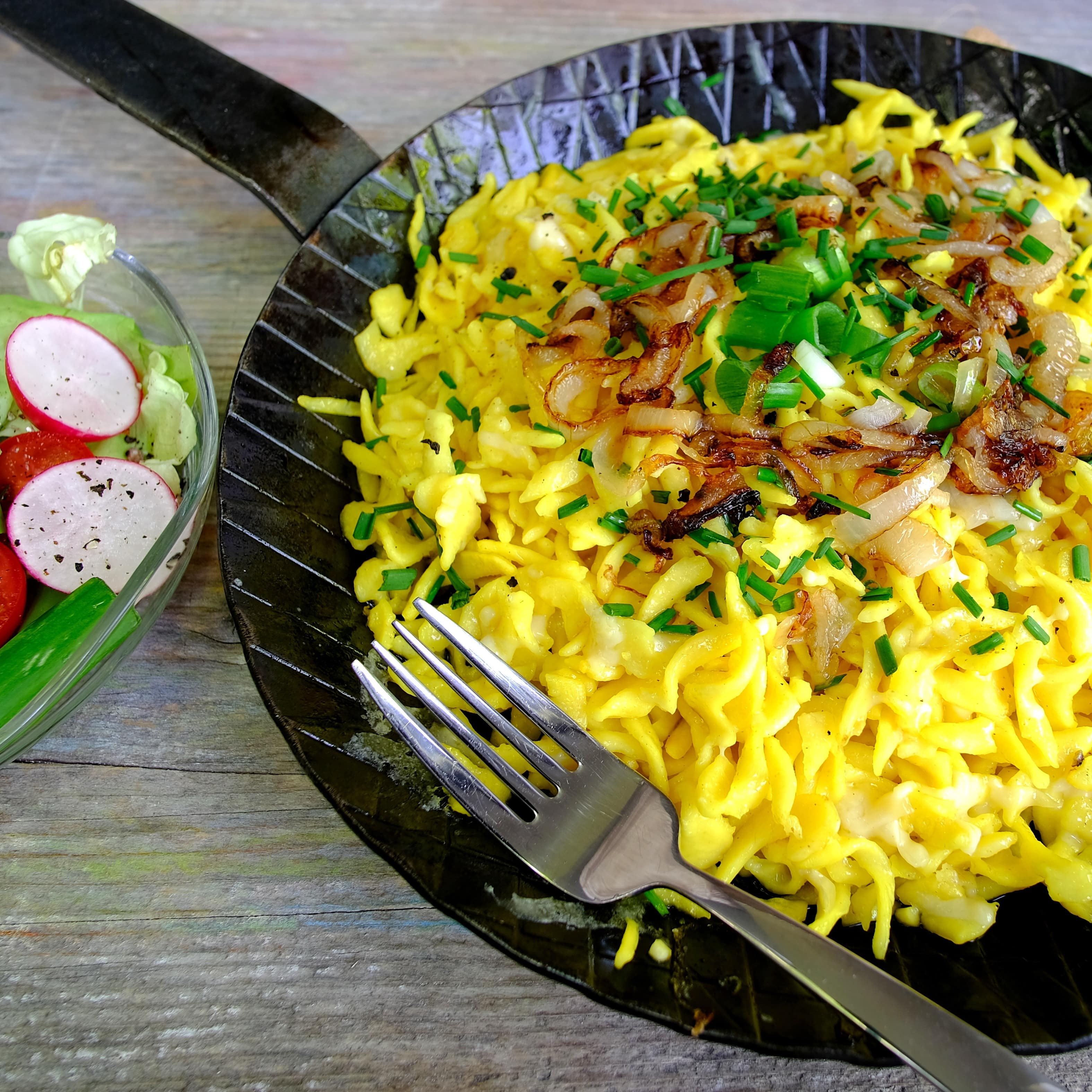 Nahaufnahme: Eine Pfanne mit Kässpätzle mit einem Salat daneben auf einem Holztisch.