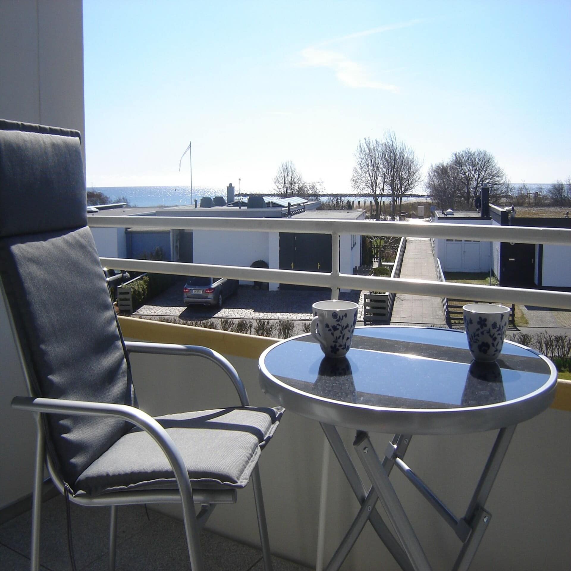 Sonniger Balkon mit Tisch und Stuhl und Blick über einiger Häuser hinweg auf das Meer.