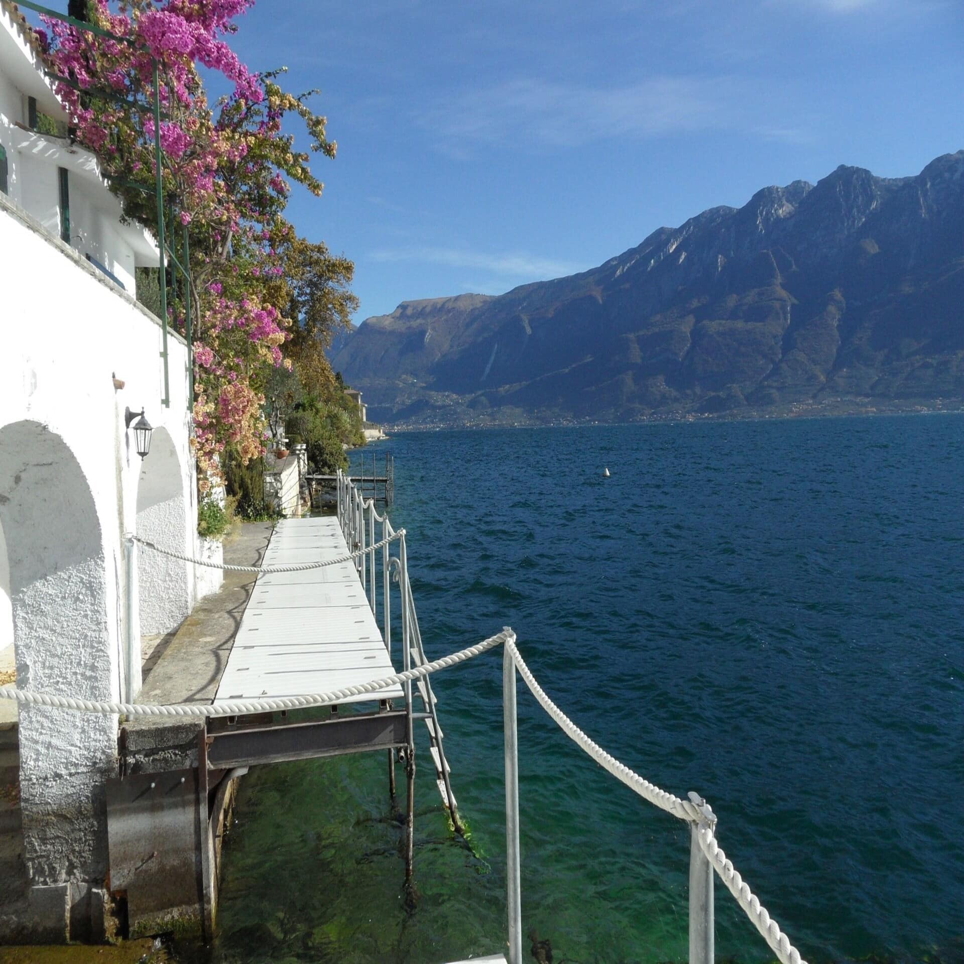 Weißes Haus mit Bootssteg direkt am Gardasee. Im Hintergrund die Berge. Die Sonne scheint.