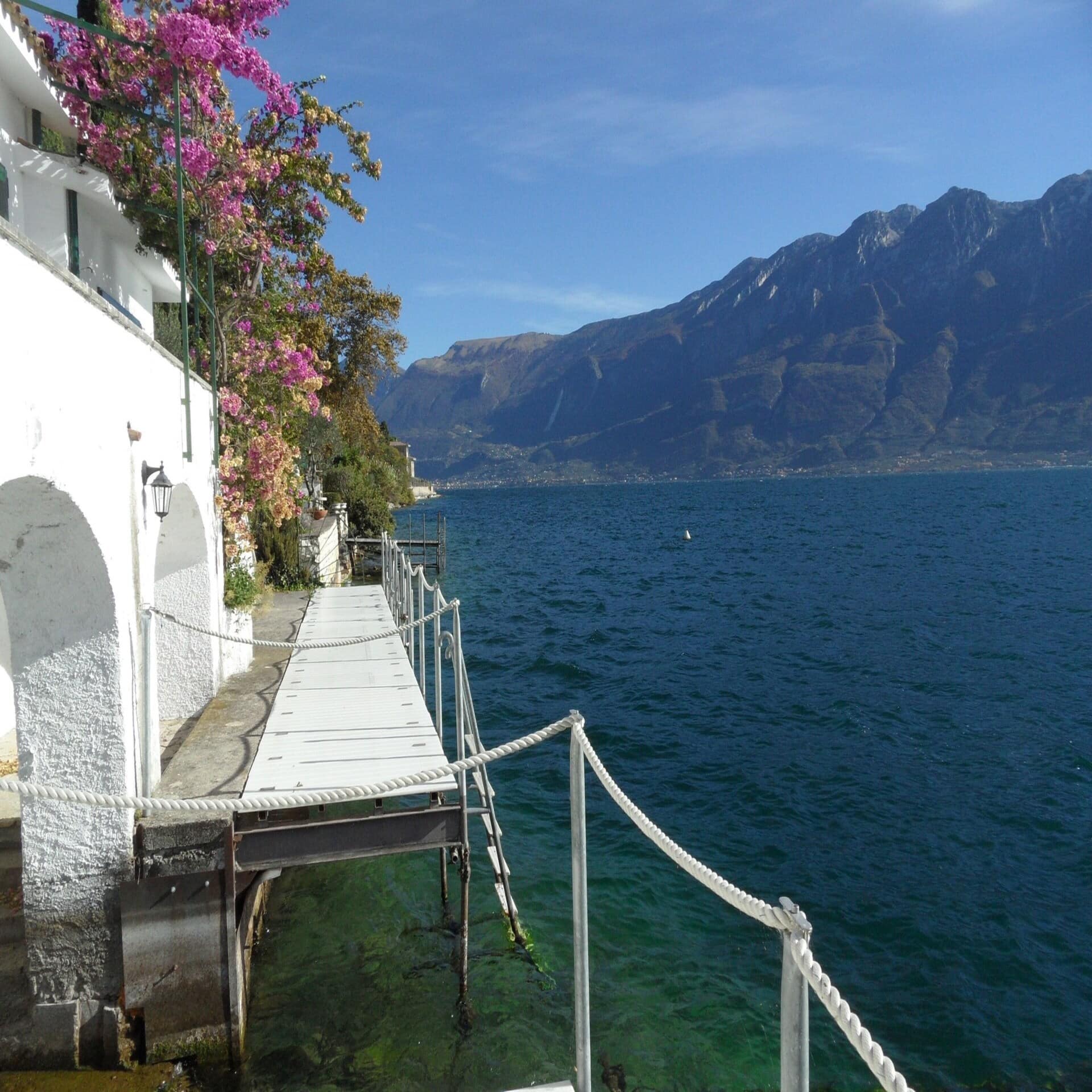 Weißes Haus mit Bootssteg direkt am Gardasee. Im Hintergrund die Berge. Die Sonne scheint. 