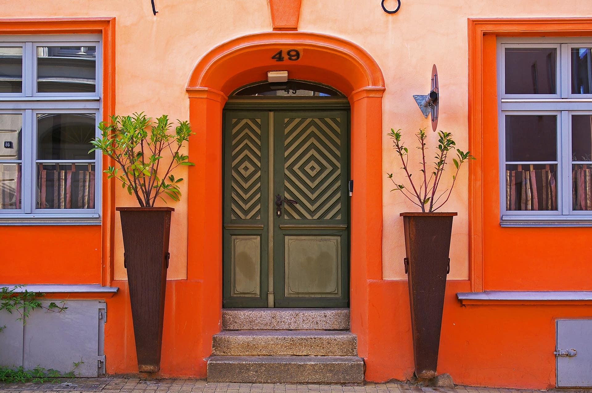 Blick auf eine orangefarbene Hausfront mit grüner Tür und blauen Fensterrahmen; daneben stehen zwei Pflanzenkübel