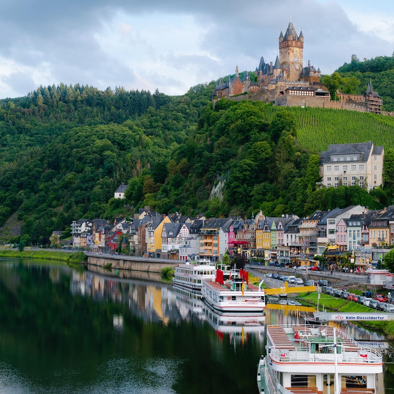 Blick auf Cochem mit Häusern am Fluss, Booten und der von Wald umgebenen Burg im Hintergrund