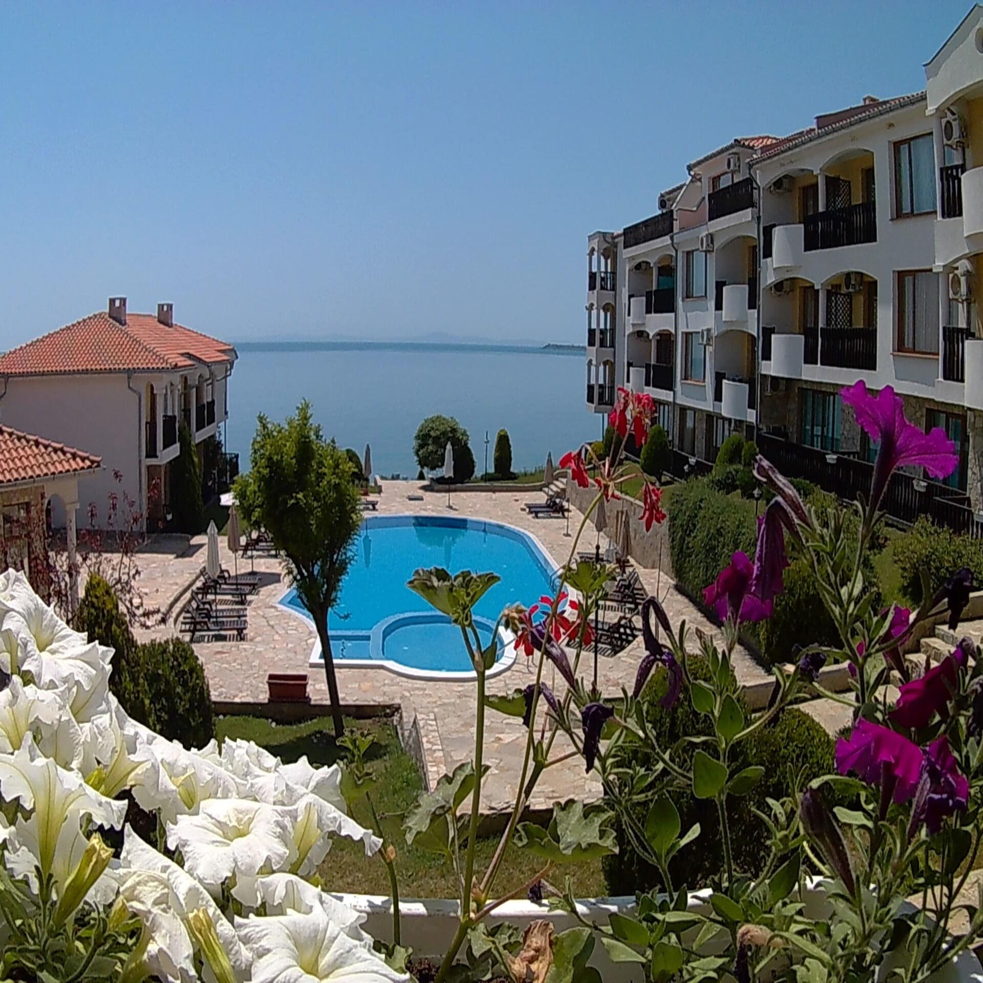 Blick von einem Balkon mit Blumen einer Wohnung in einer Ferienanlage auf den Poolbereich und das Meer. 