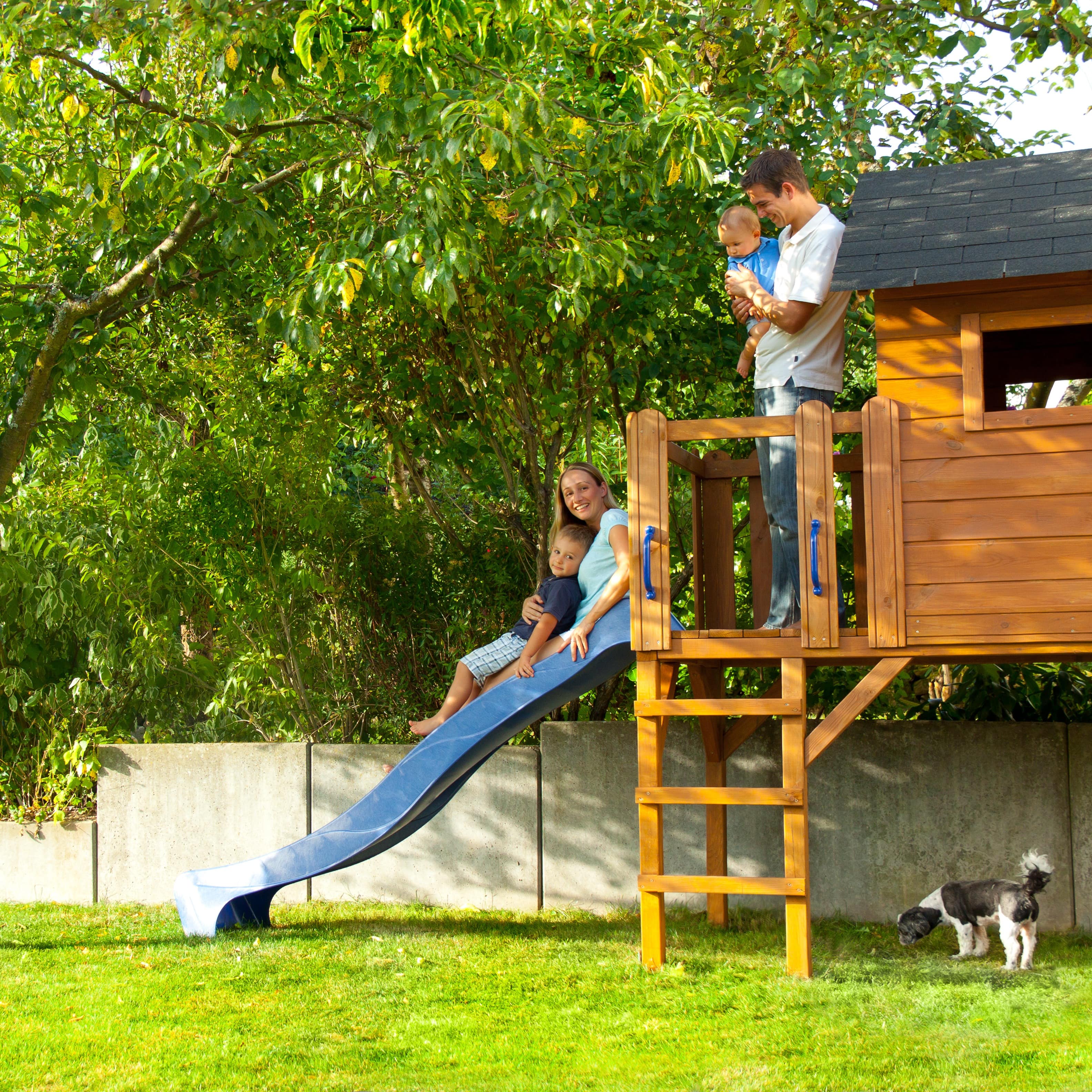 Familie mit kleinem Jungen und Baby auf einem Spielhaus mit Rutsche im Garten. Ein Hund ist zu sehen.
