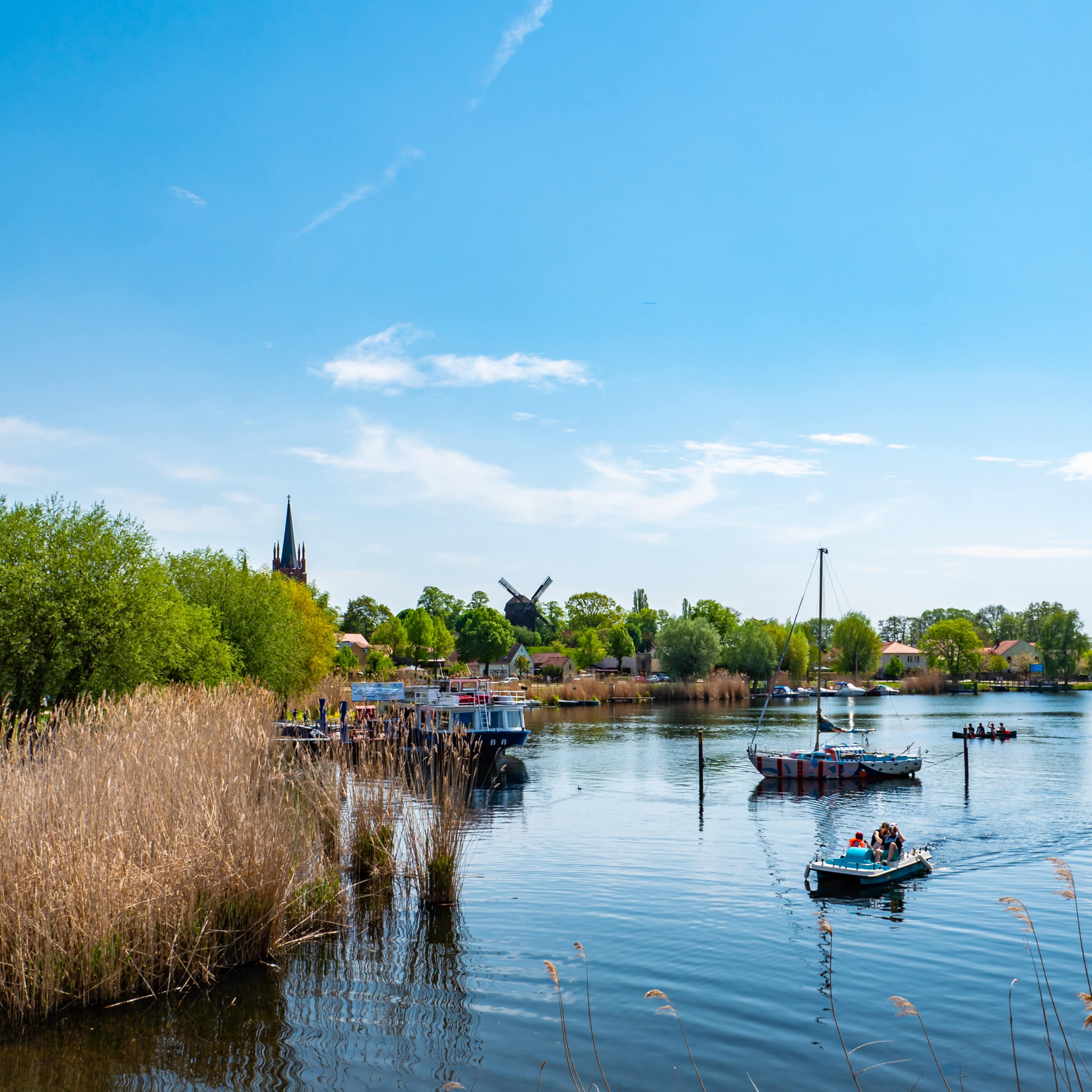 Blick über den kleinen Hafen von Werder, einige kleine Boote schippern auf dem Wasser, im Hintergrund Kirche und Windmühle.