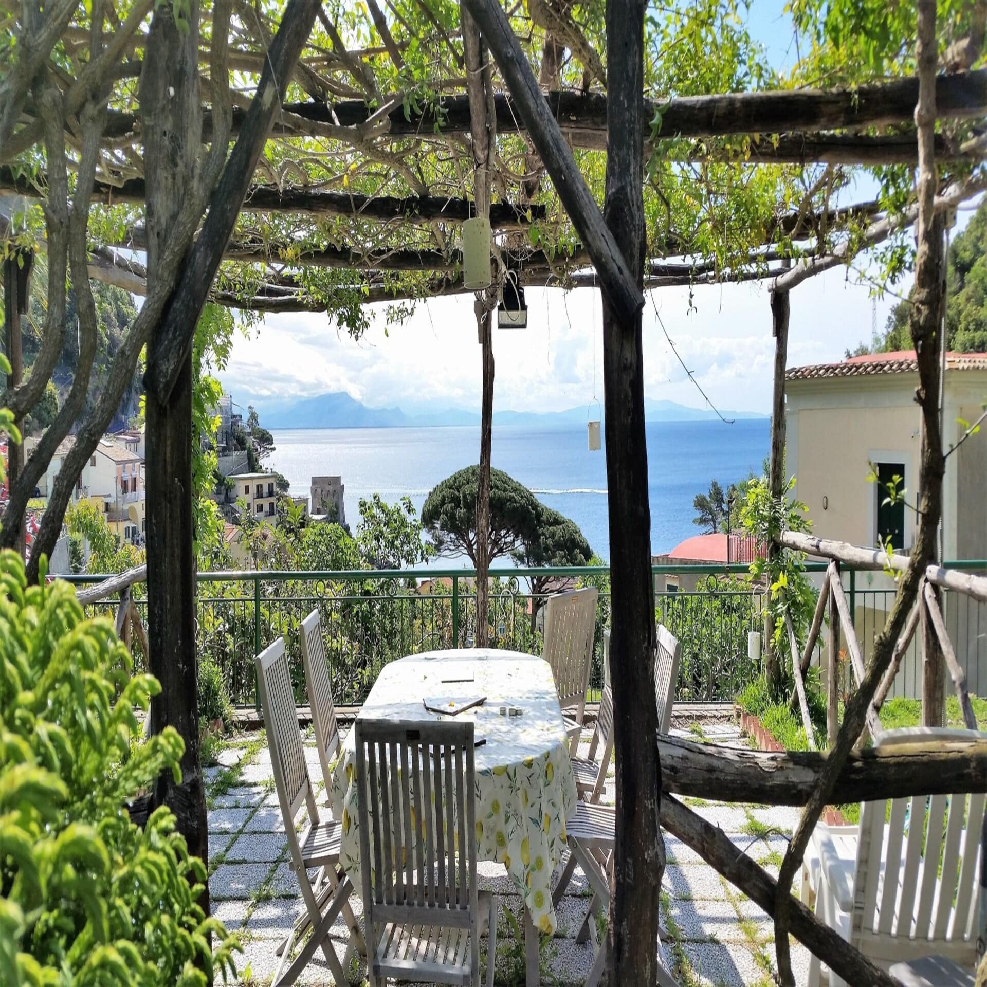 Eine mit Weinranken überwucherte Terrasse mit Tisch und Stühlen und Bick auf einige Häuser und einen See.