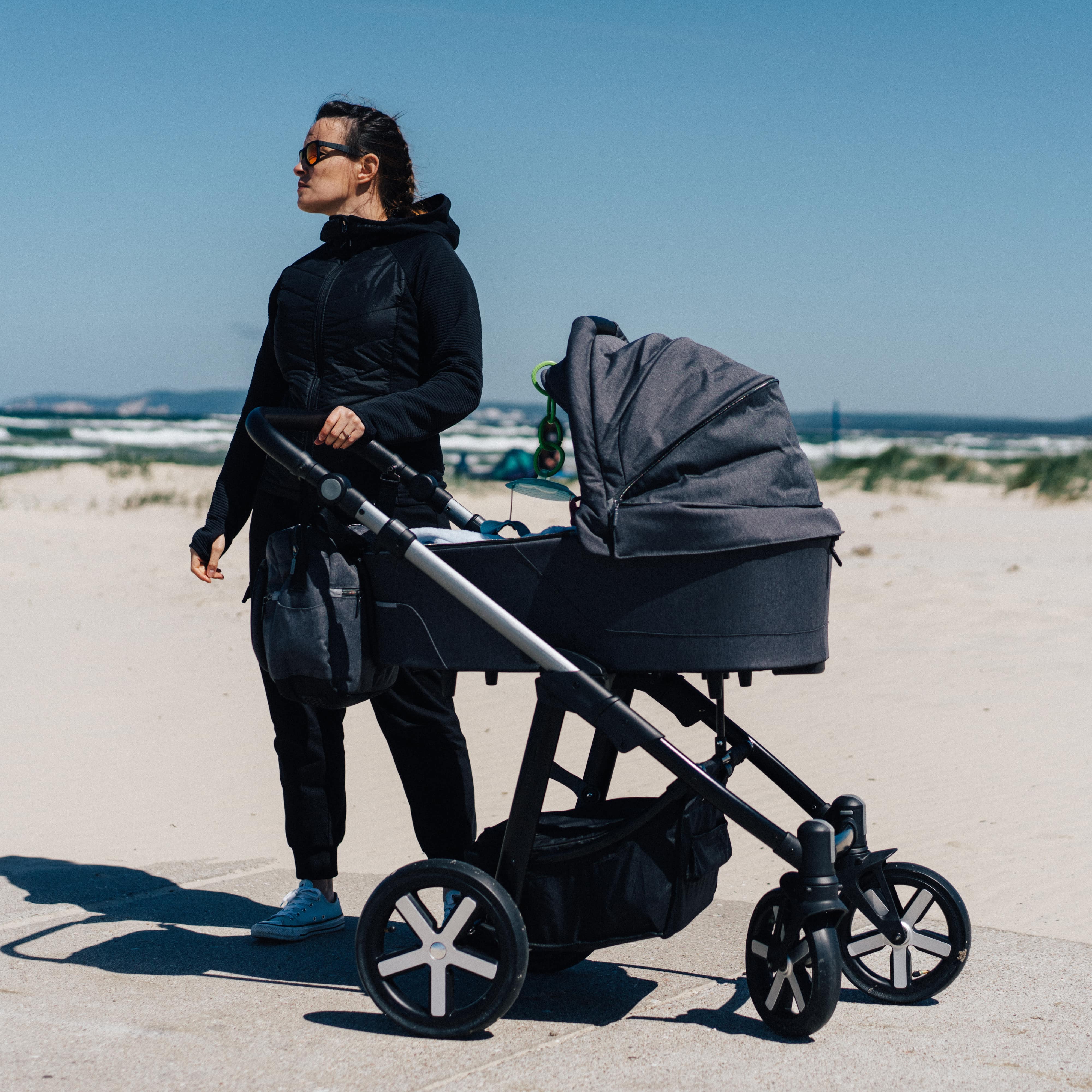 Mutter in schwarzer Kleidung mit Kinderwagen am Strand.