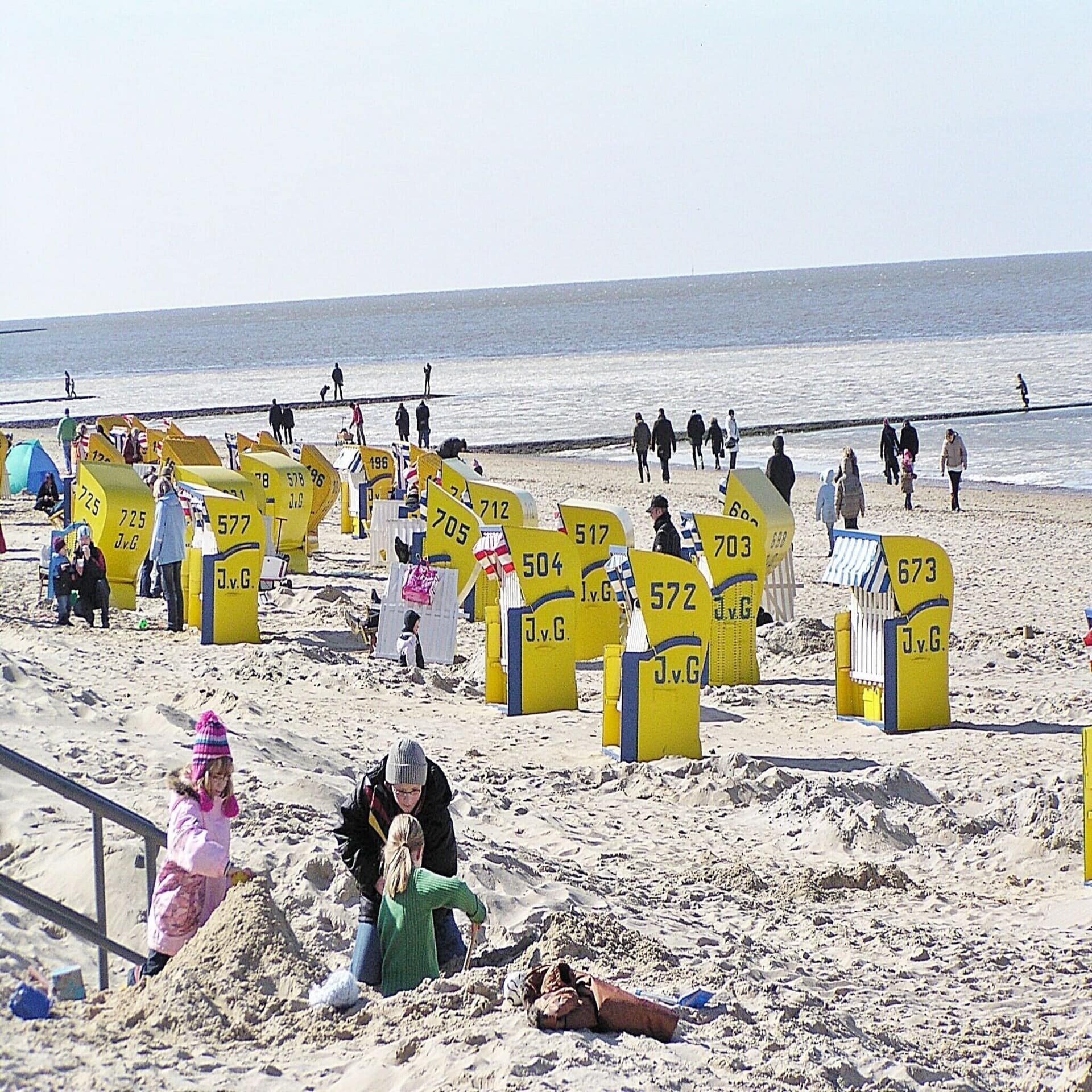2 Kinder spielen mit einer Frau am Strand. Es sind gelbe Strandkörbe und herbstlich gekleidete Leute zu sehen. 