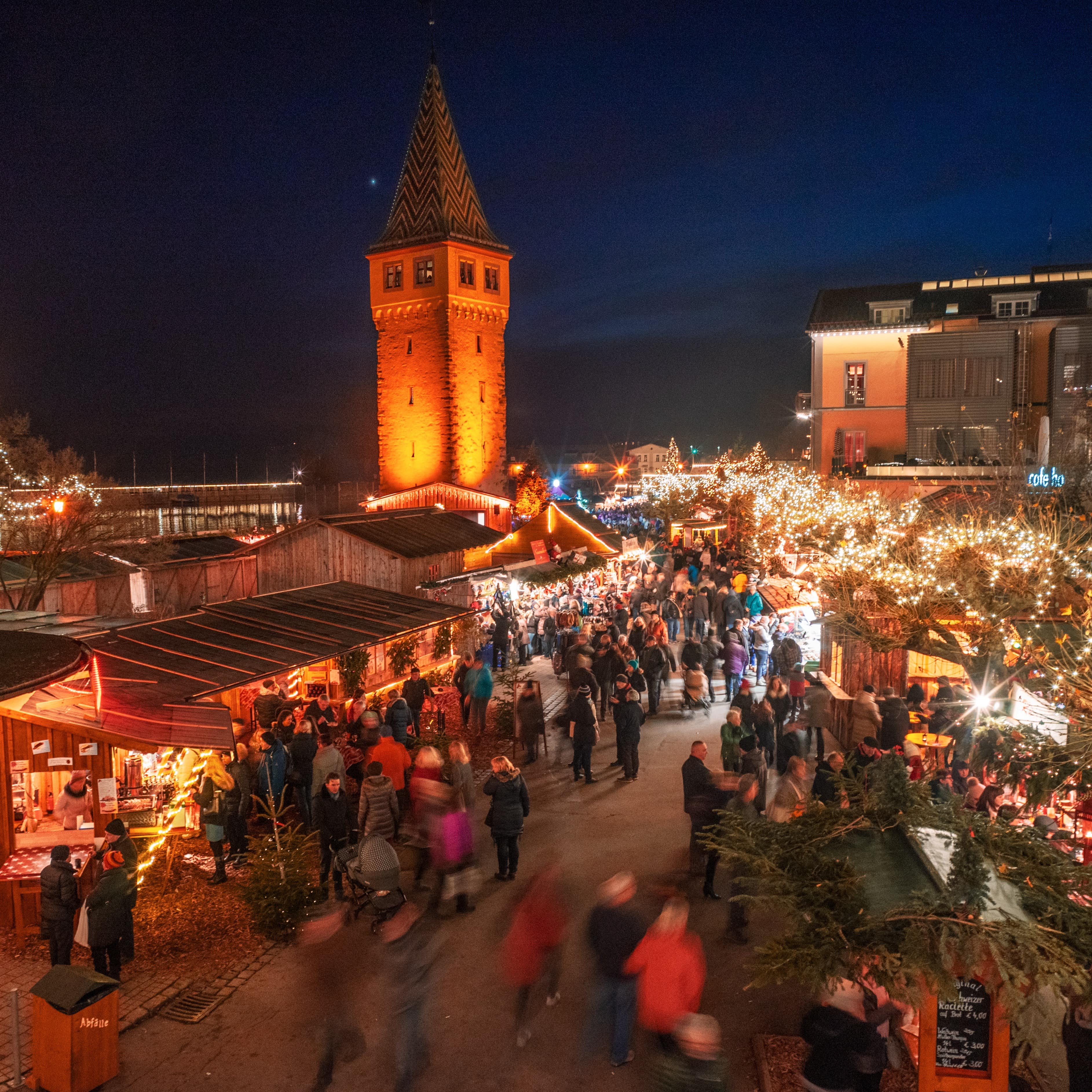 Hell erleuchteter Weihnachtsmarkt am Lindauer Hafen am Abend. Viele Leute laufen an den Ständen vorbei.