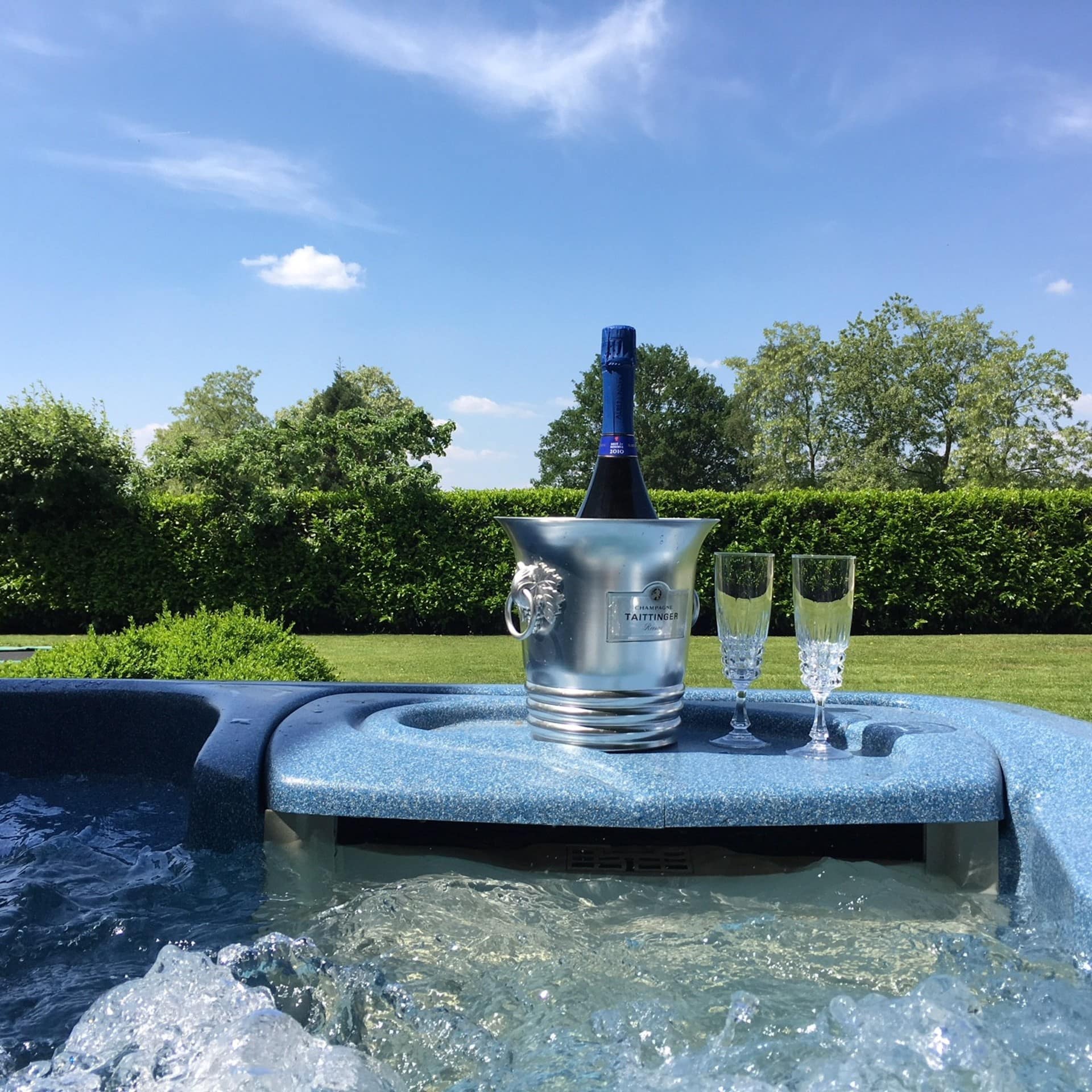 Blick von einem Whirlpool in einem grünen Garten auf Champagner, Gläser und den Garten.