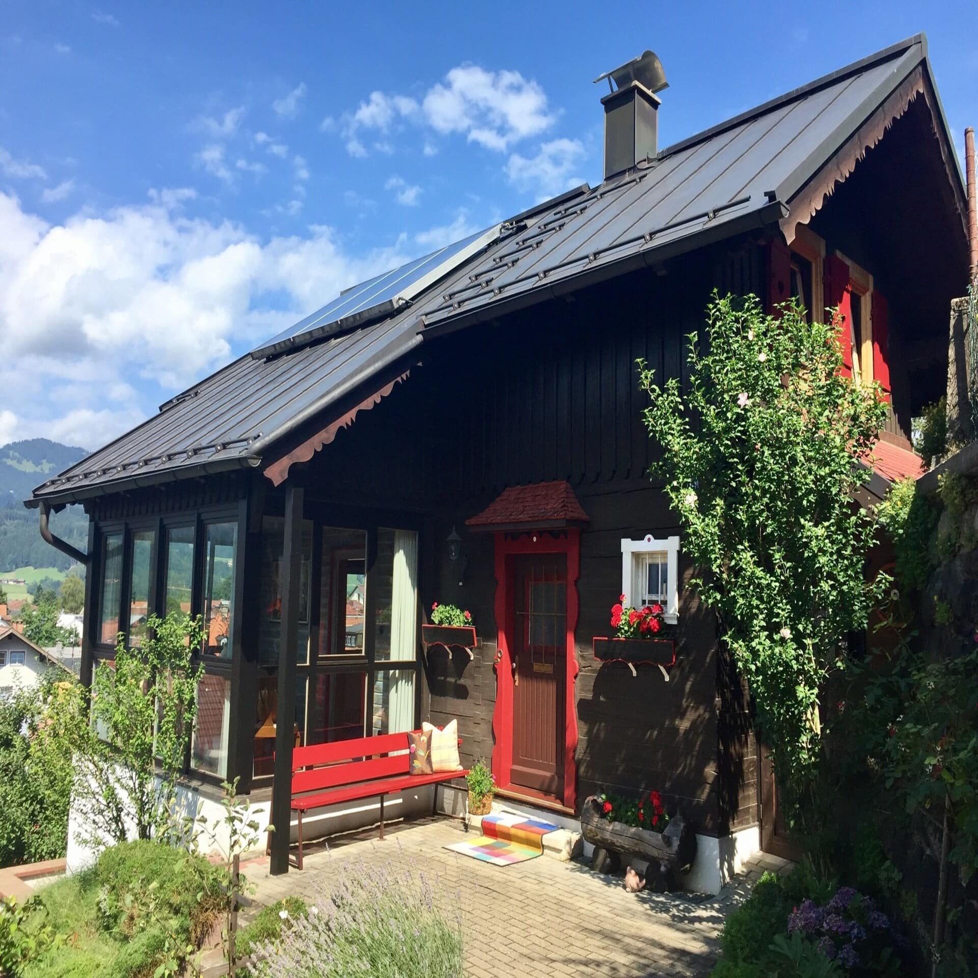 Kleines braunes Holzhaus mit roter Sitzbank und verglastem Wintergarten mit Blick auf den Ort. 