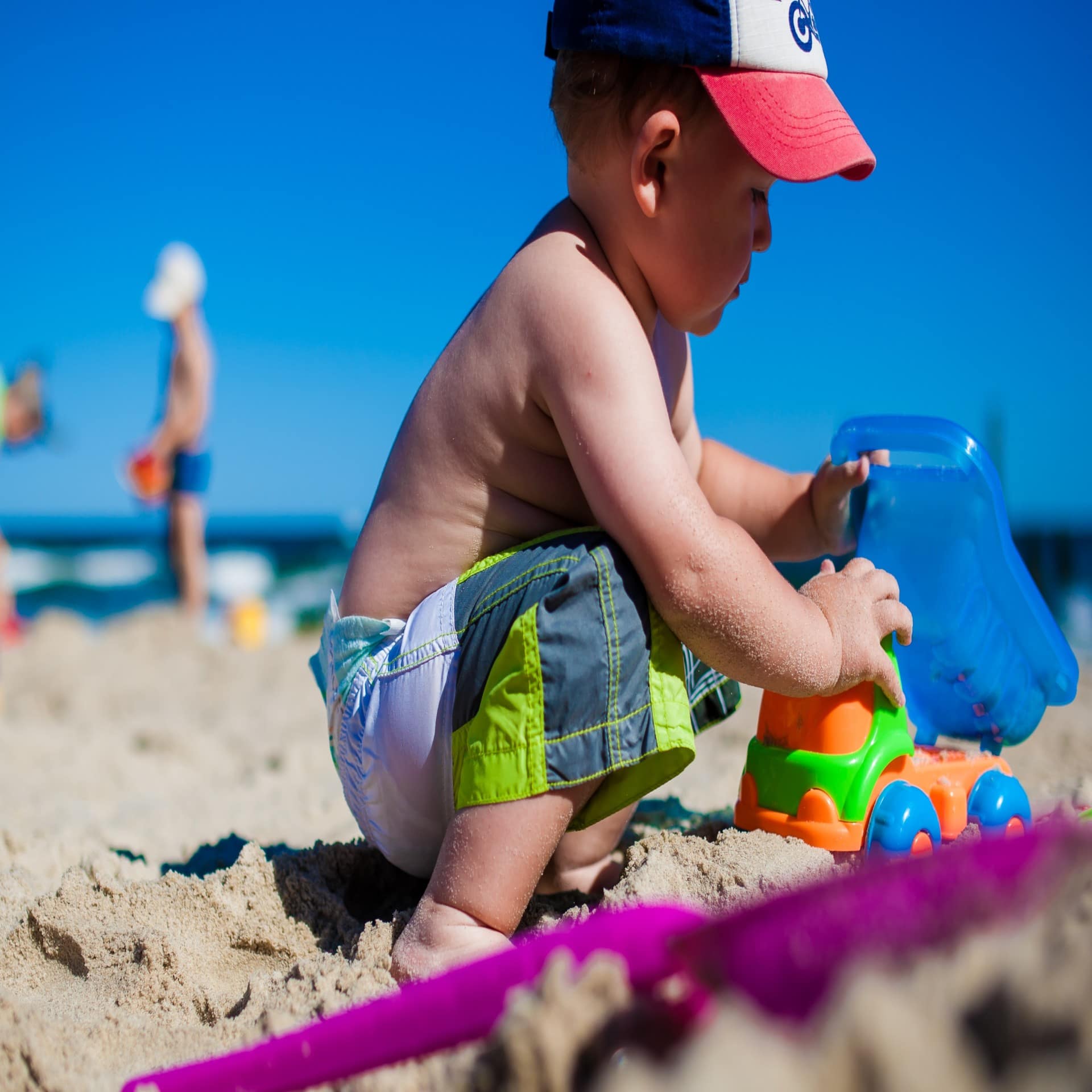 Nahaufnahme: Ein kleiner Junge mit Badehose und Mütze spielt mit einem kleinen Lastwagen am Strand. Die Sonne scheint. 