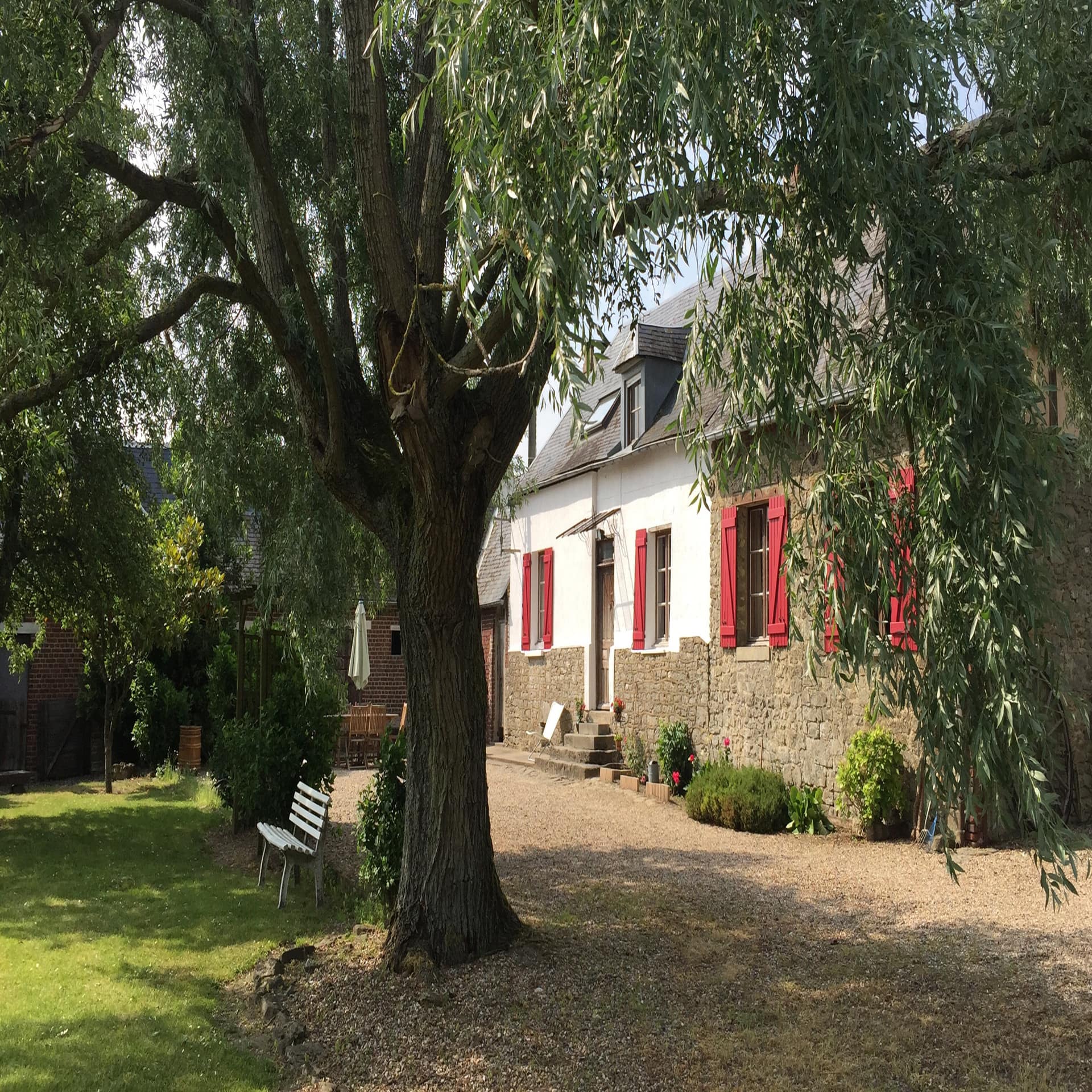 Bauernhaus mit roten Fensterläden, davor ein Garten mit Baum und Bank.