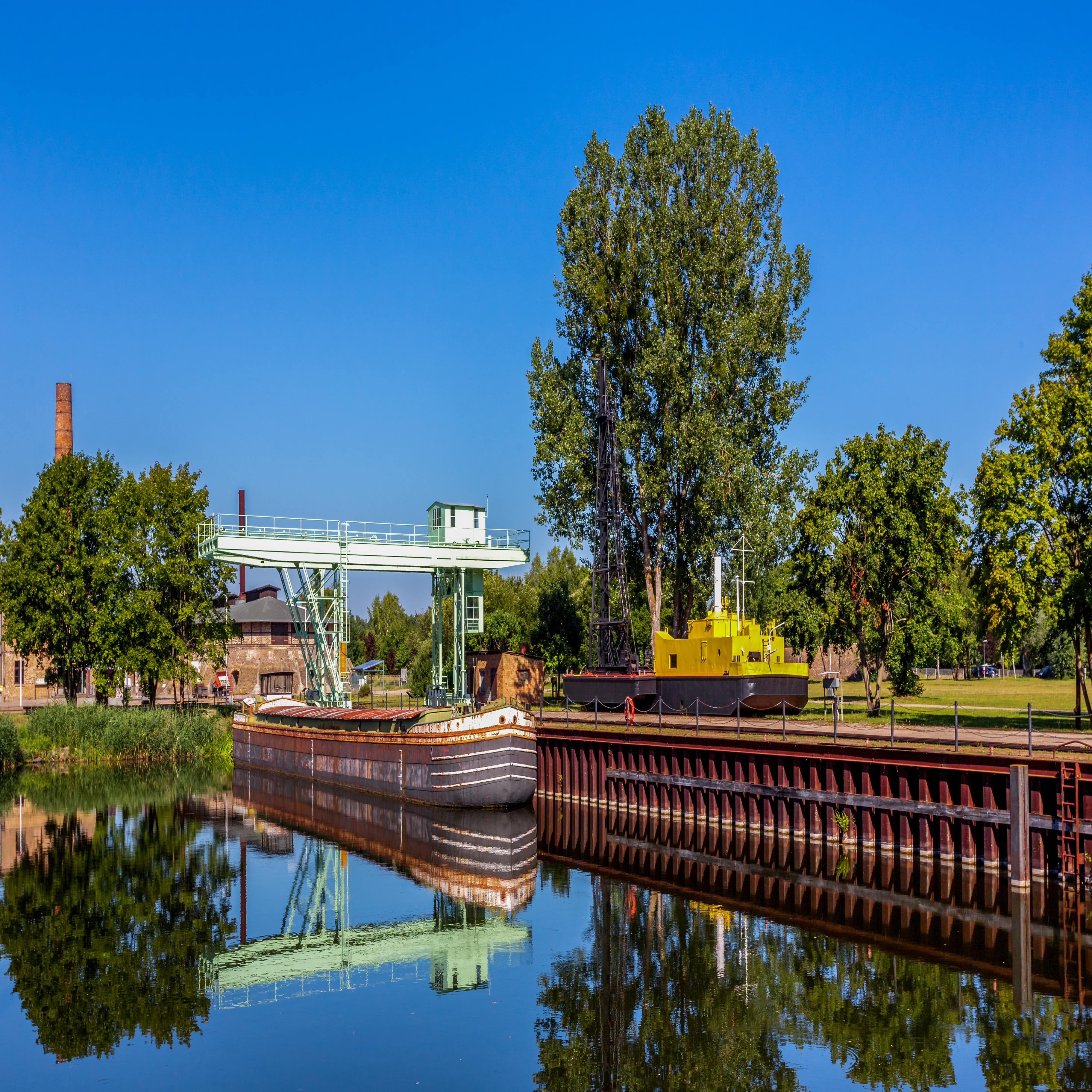 Blick von außen auf die Anlage des Ziegeleiparks. Ein rostiger Kahn im Wasser, dahinter ein Kran. Rechts ein gelb-schwarzes Fahrzeug.