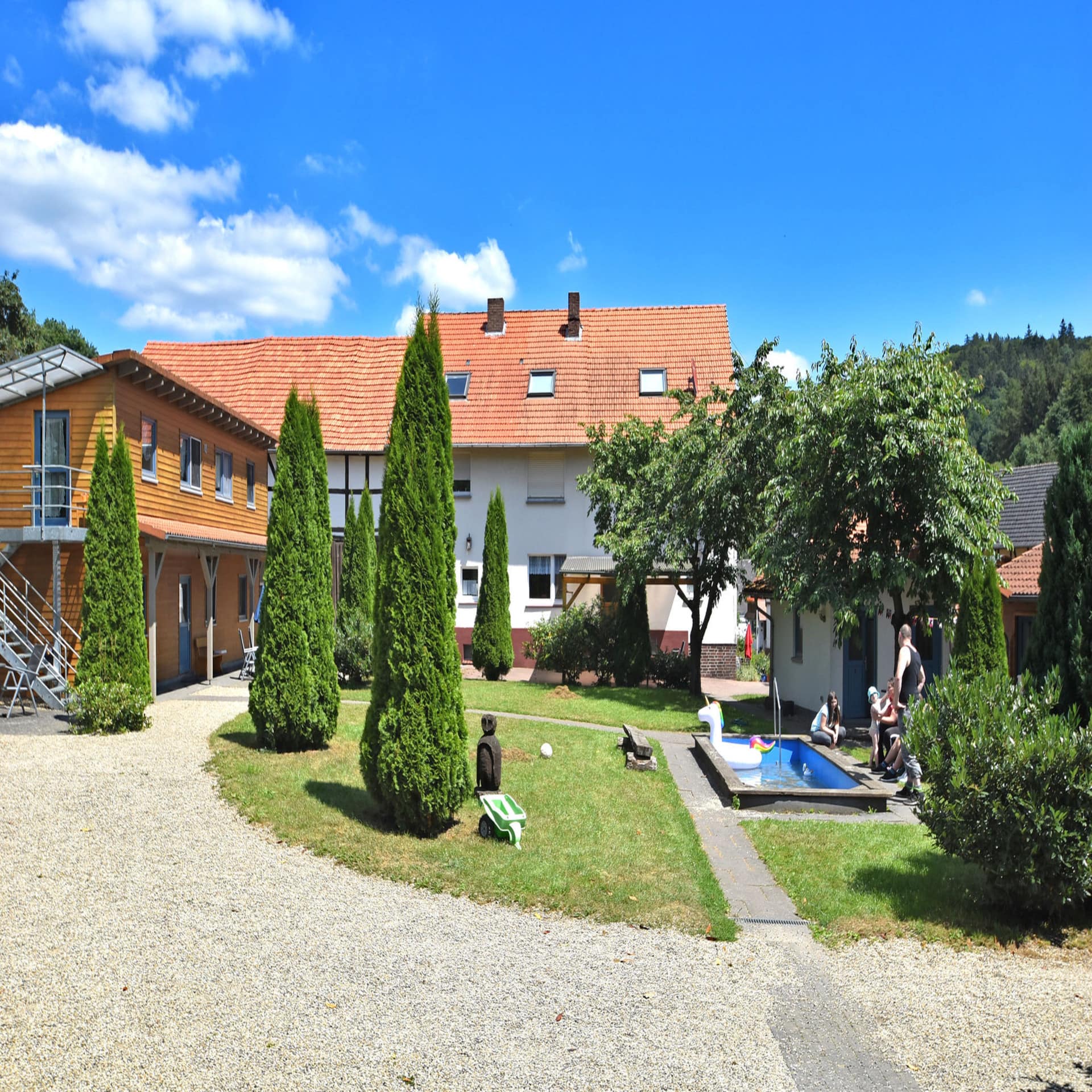 Bauernhof mit Garten in dem ein Pool steht. Ein großes Schwimmtier im Pool, eine Familie mit Kleinkind daneben.