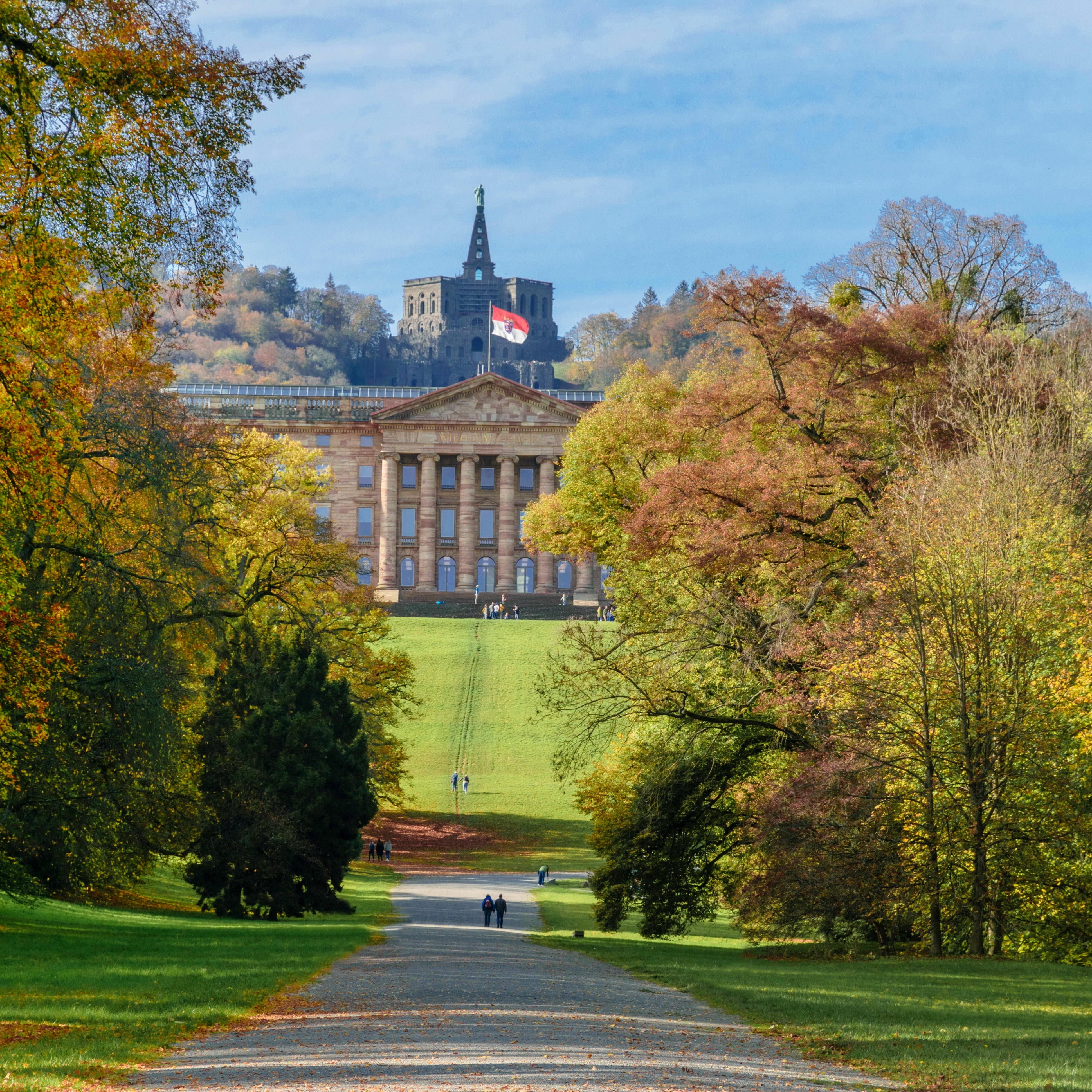 Sonniger Herbsttag: Blick entlang eines Spazierweges zwischen Bäumen auf Schloss Wilhelmshöhe und den Herkules dahinter.
