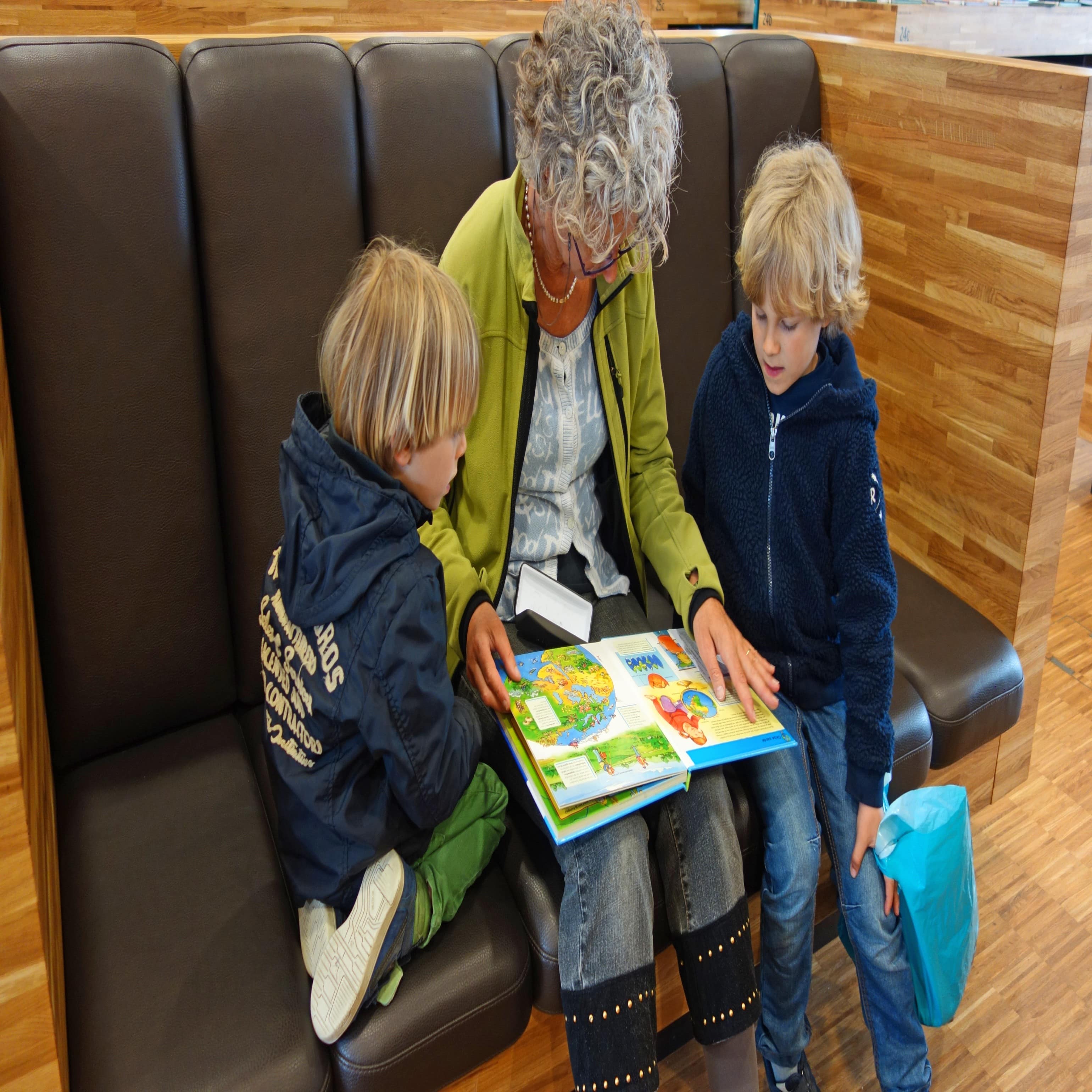 2 Jungen sitzen rechts und links neben ihrer Großmutter aus einer Bank und lesen ein Buch.