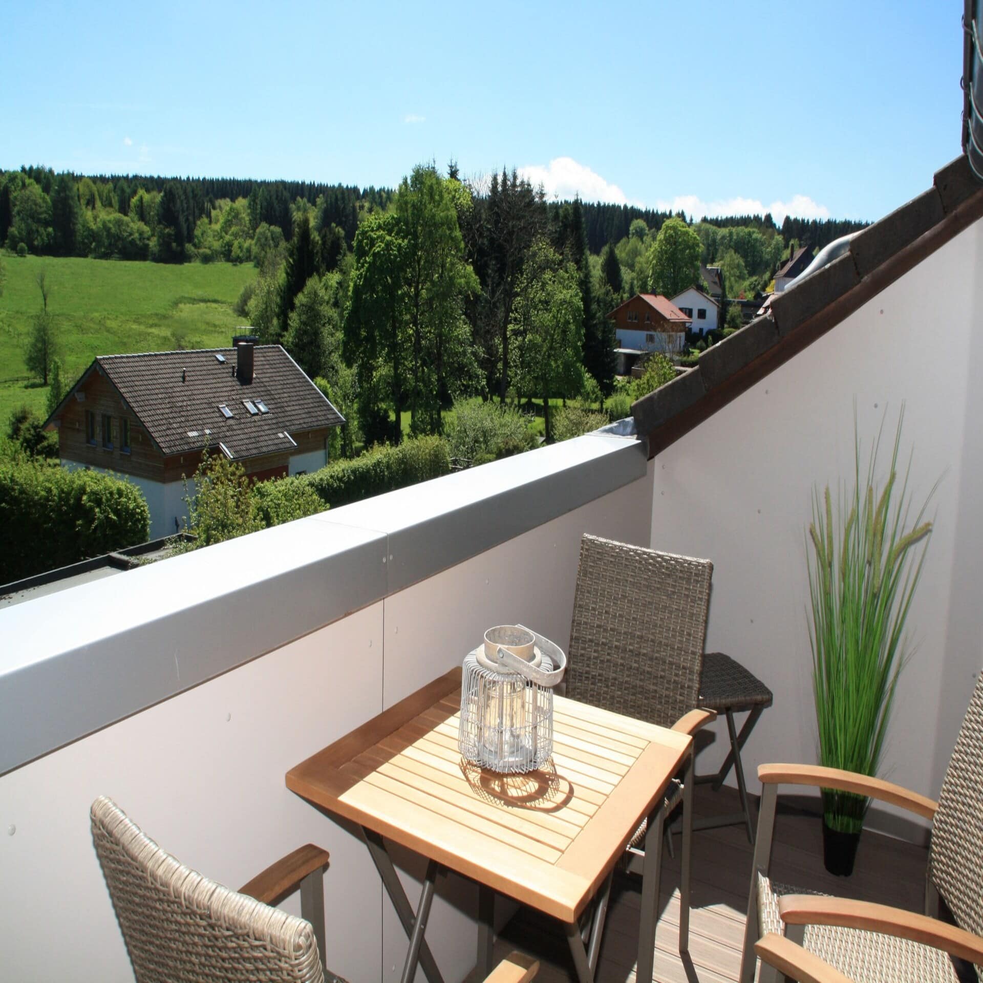 Balkon mit Tisch und 4 Stühlen und Blick auf einige Häuser und das grüne Umland. Die Sonne scheint.