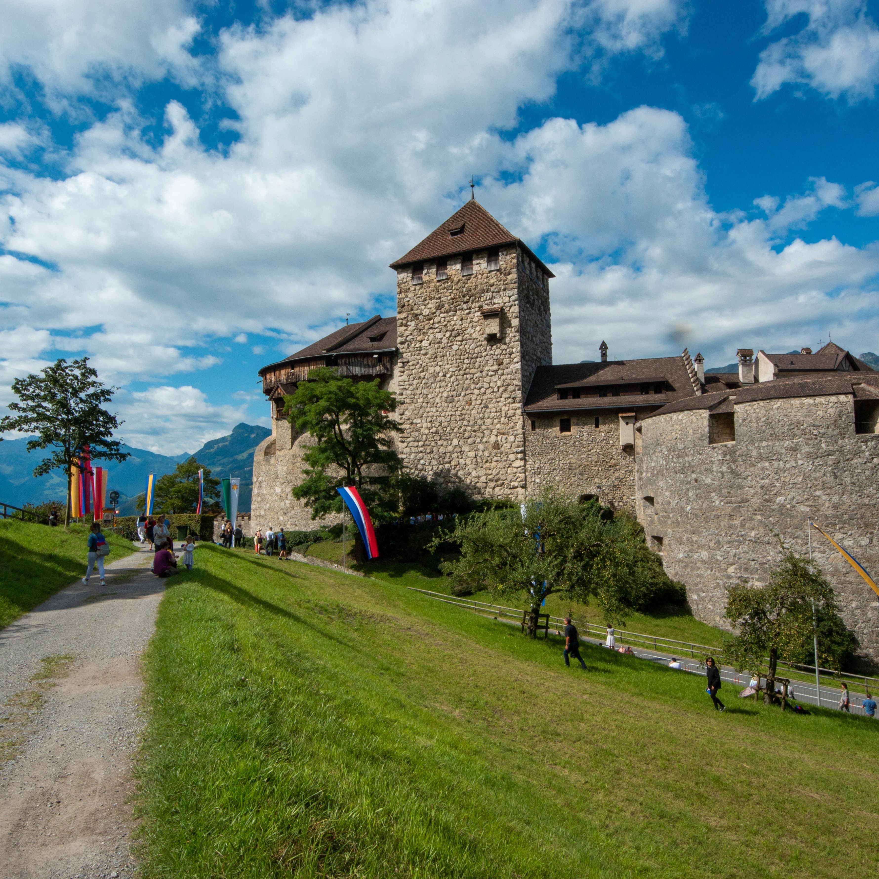 Ein Schotterweg mit Fahnen führt zu Schloss Vaduz, Personen laufen auf das Schloss zu.