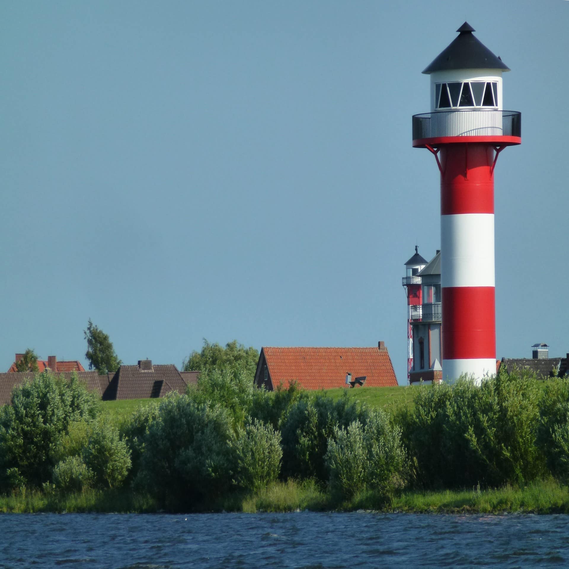 Blick über das Wasser auf einen rot-weiß gestreiften Leuchtturm und Häuserdächer hinter dem Deich. 