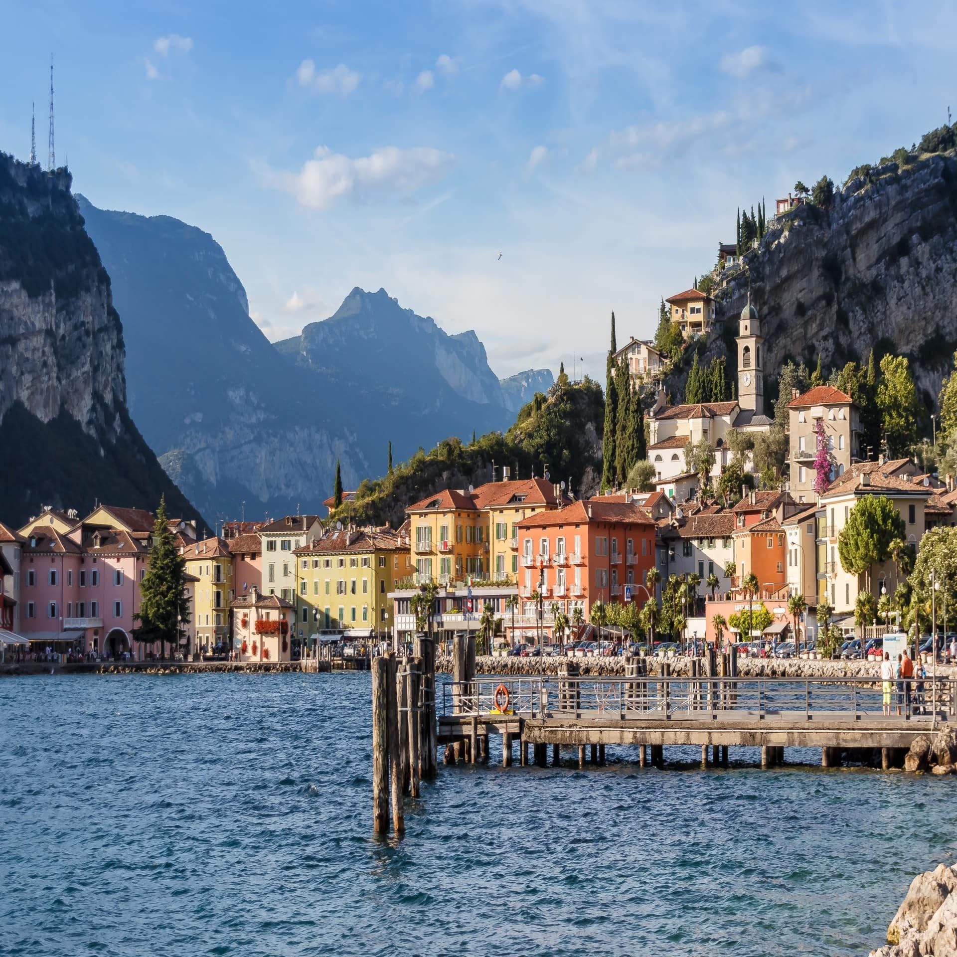 Blick vom Gardasee auf eine Stadt am Ufer mit Bergen im Hintergrund. 