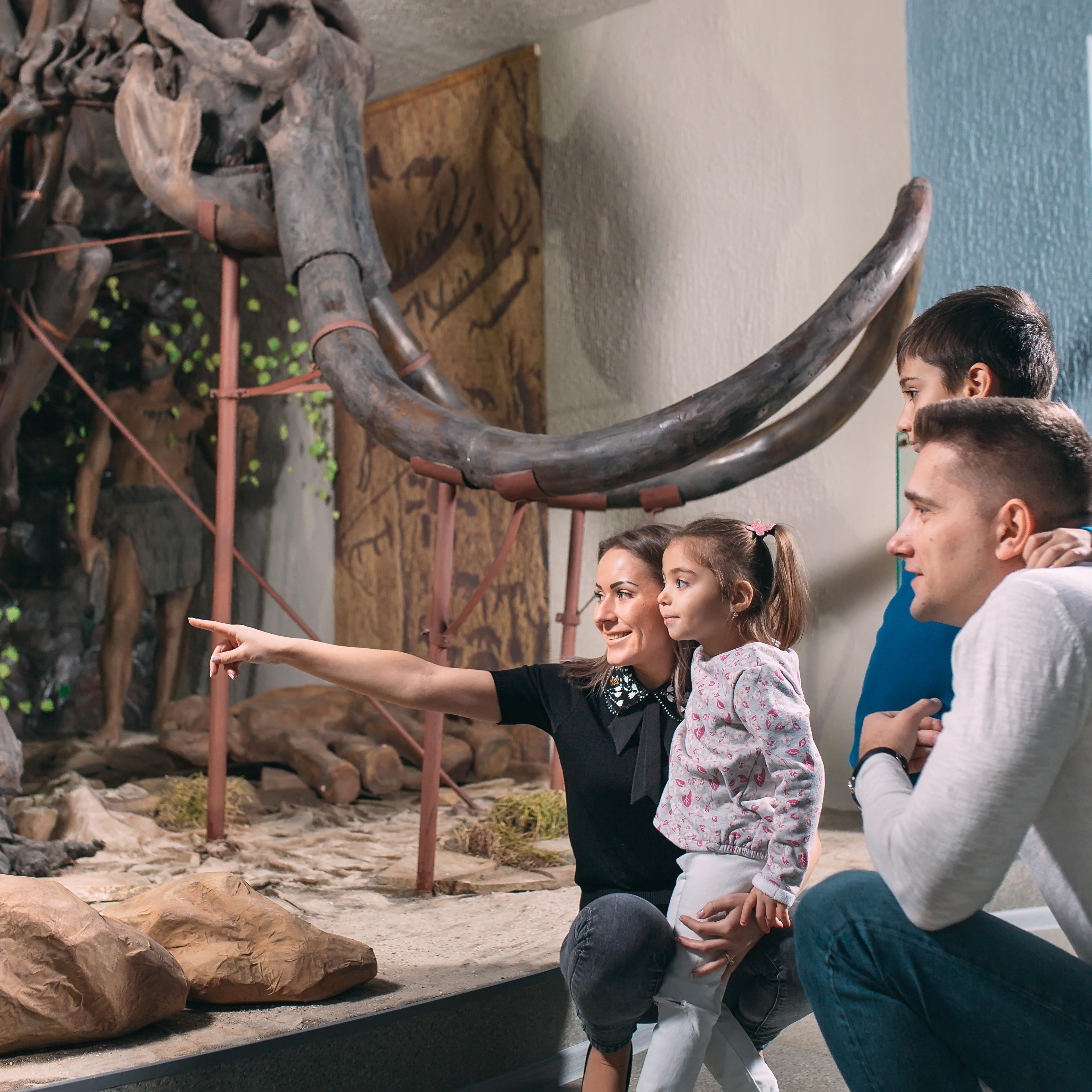 Familie mit 2 Kindern hockt vor einem Mammutskelett und schaut etwas an. Die Mutter zeigt auf etwas.