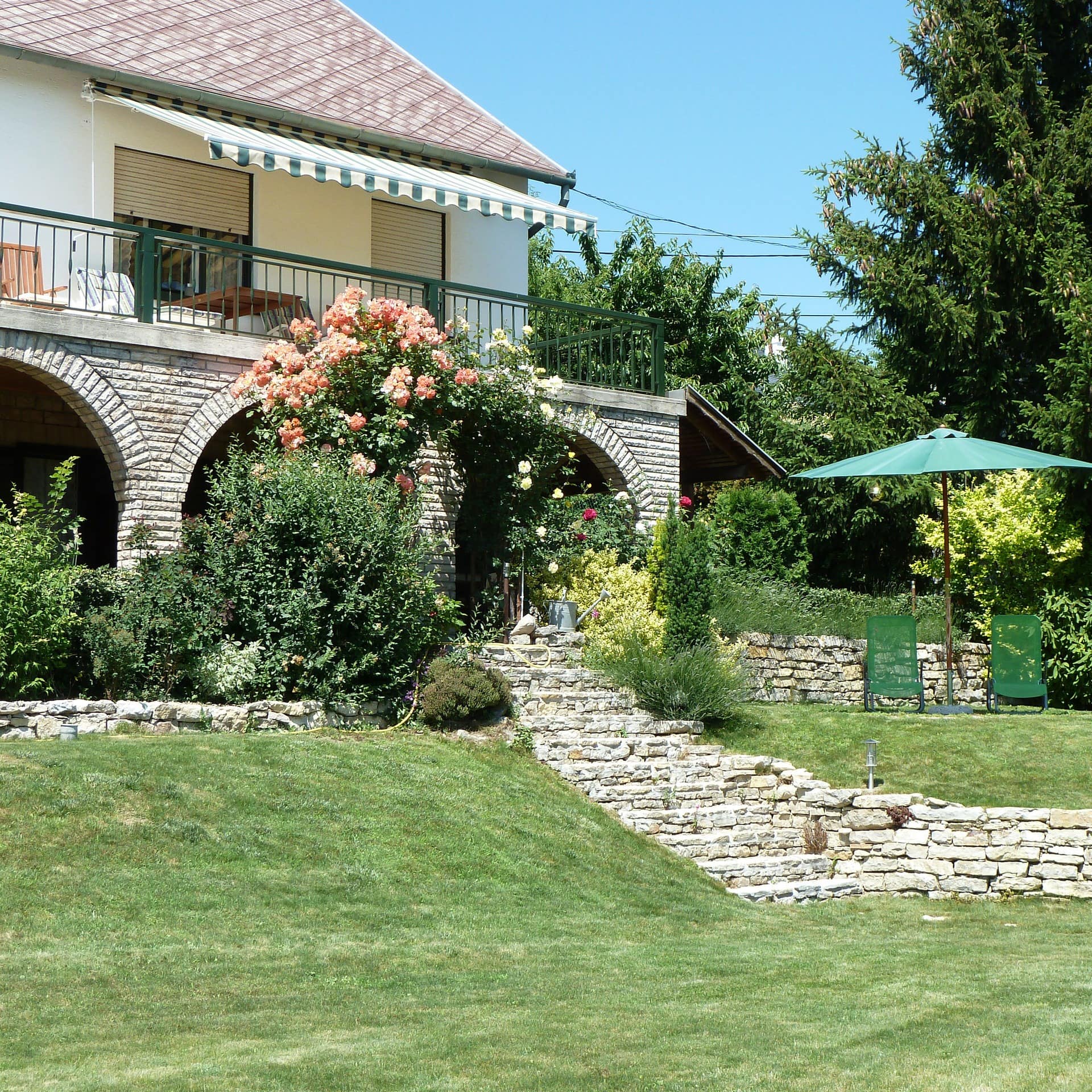 Ein Ferienhaus am Balaton mit Balkon und Rundbögen und einem großen Garten mit Stühlen und Sonnenschirm