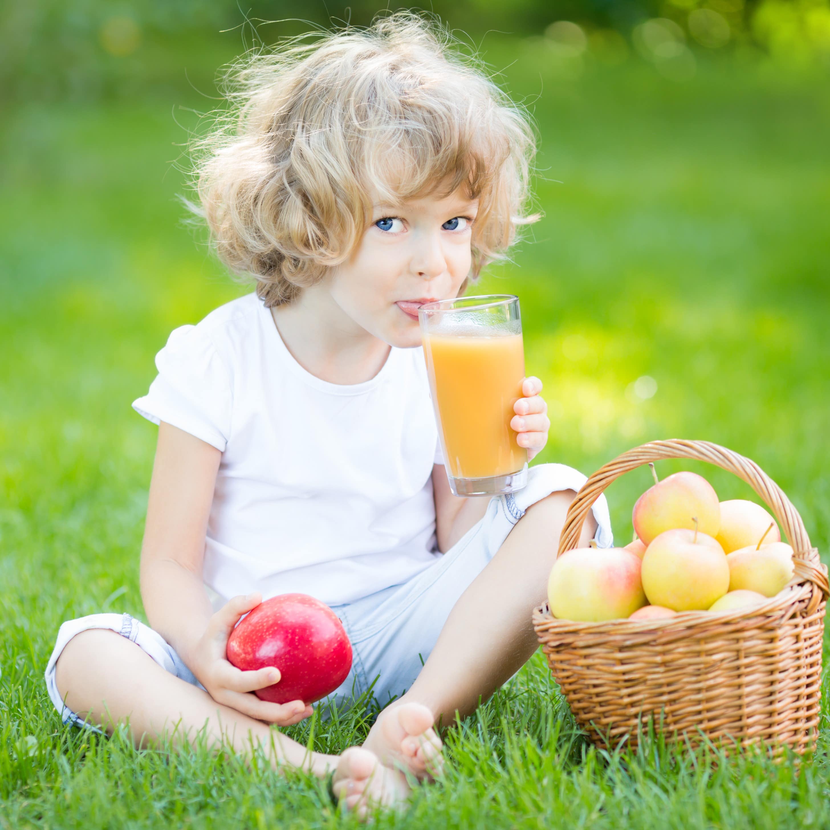 Ein Kind auf einer Wiese mit einem Glas Apfelsaft und einem Apfel in den Händen. Daneben ein Korb Äpfel.