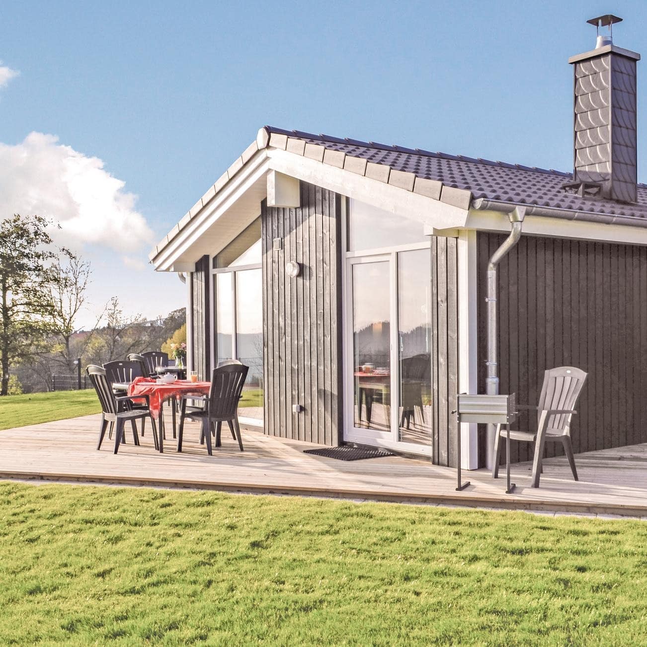 Blick auf ein einstöckiges, großes Ferienhaus mit Terrasse und Sitzgelegenheiten, umgeben von Natur