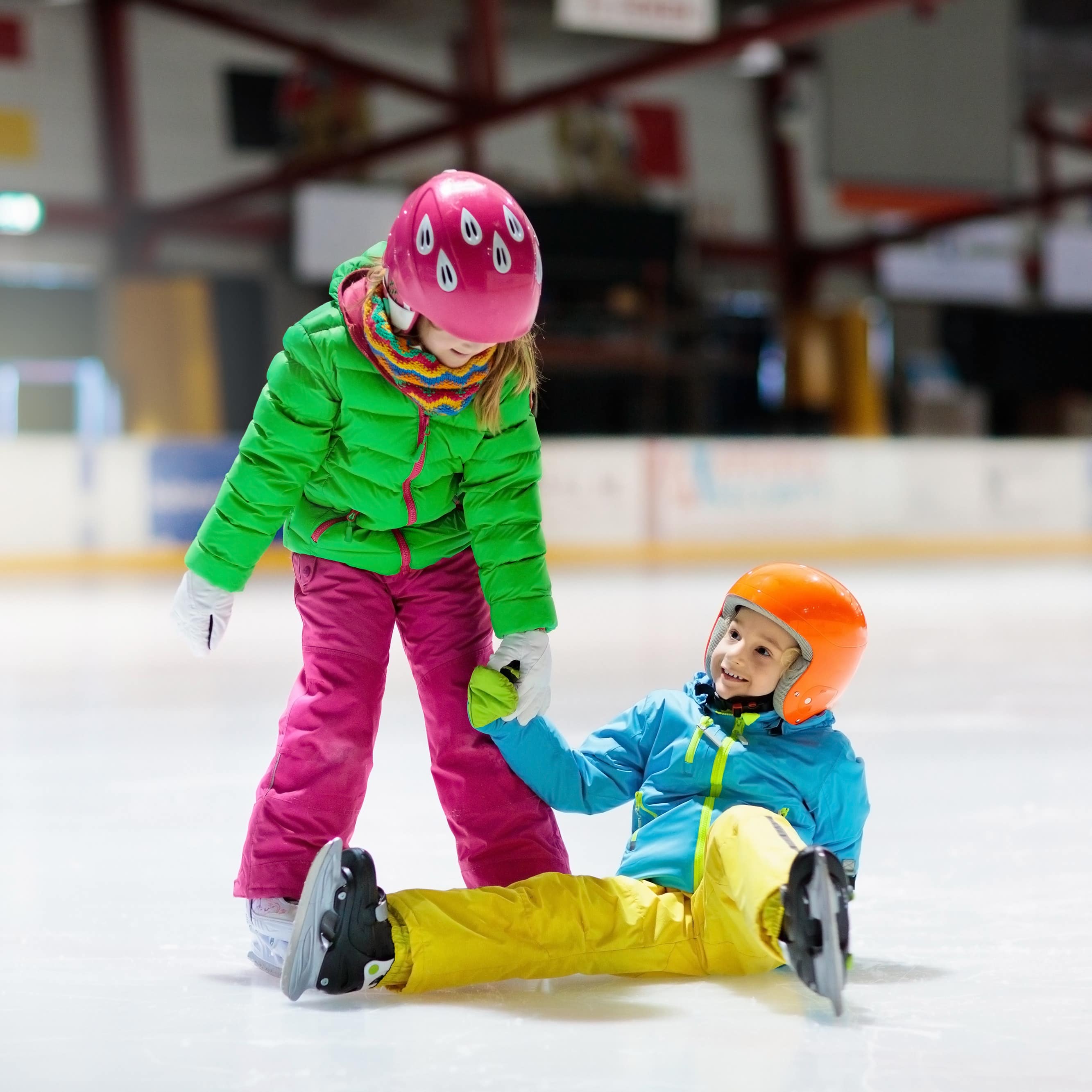 2 Kinder in bunter Winterkleidung beim Schlittschuhlaufen. Junge sitzt auf dem Boden, Mädchen hilft ihm auf.