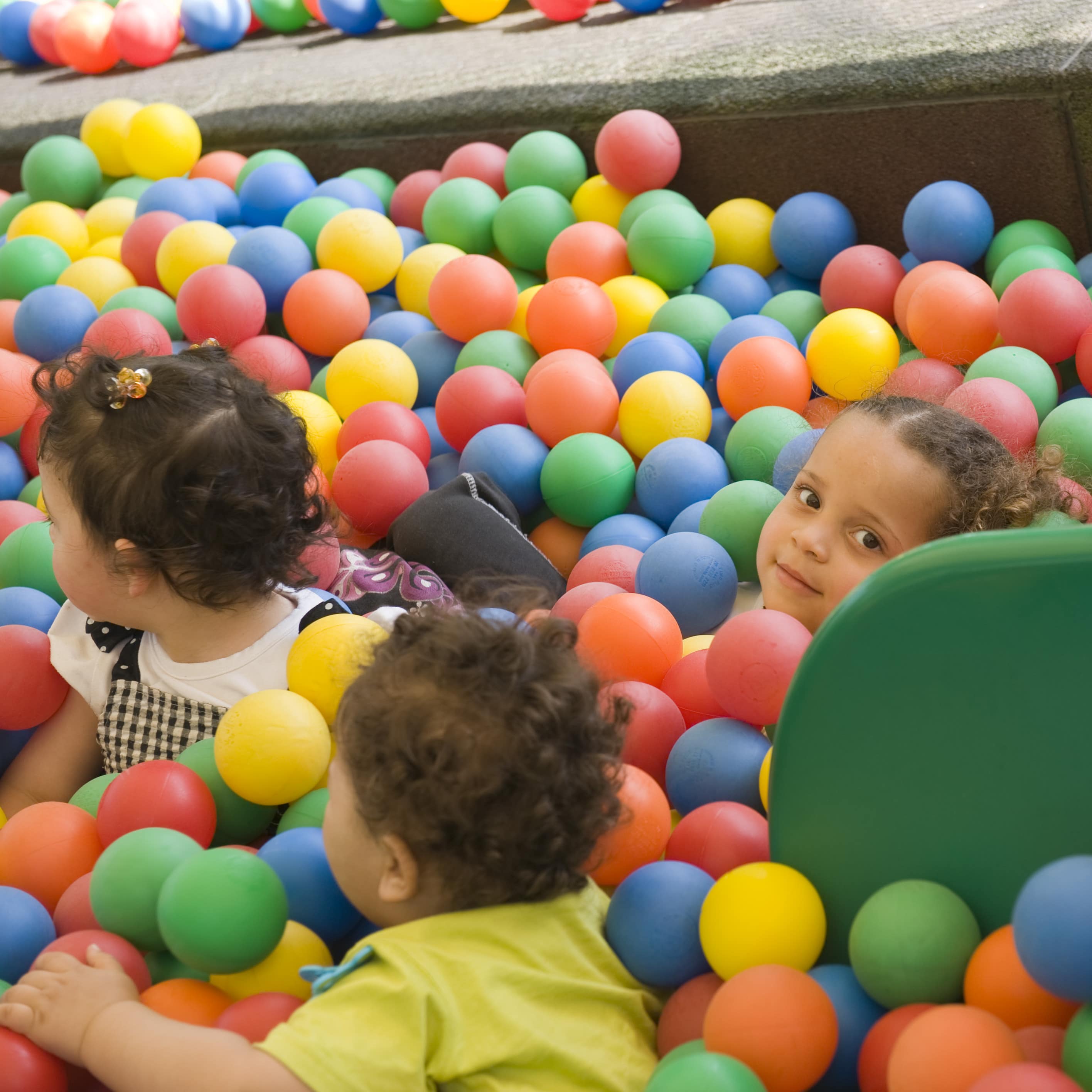 3 kleine Kinder in einem Bällebad, ein Mädchen sitzt vor einer Rutsche und schaut in die Kamera.