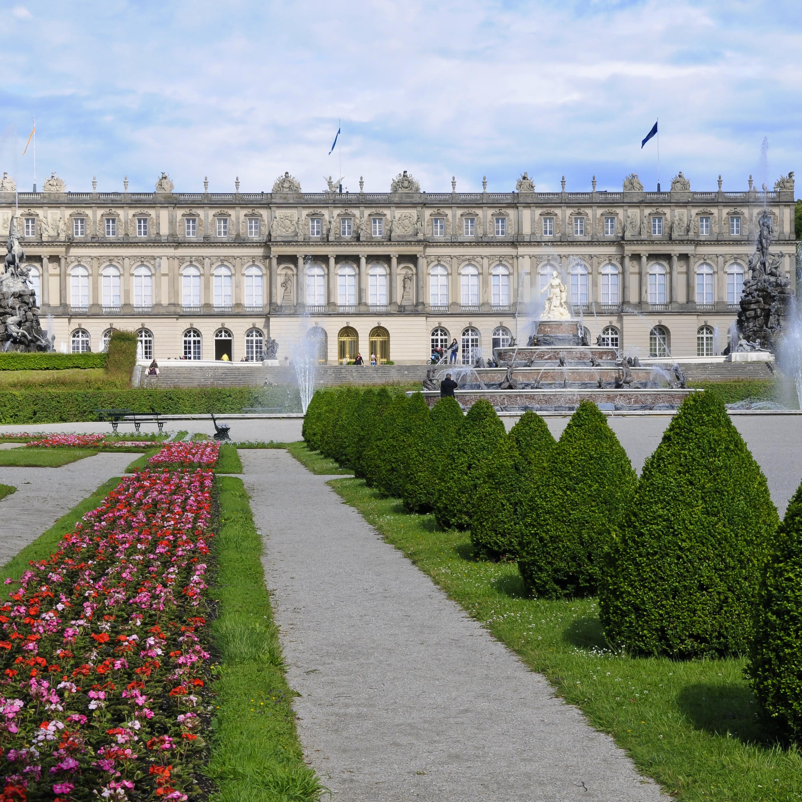 Blick auf das Schloss Herrenchiemsee, davor ein Brunnen mit Fontänen und ein Garten.