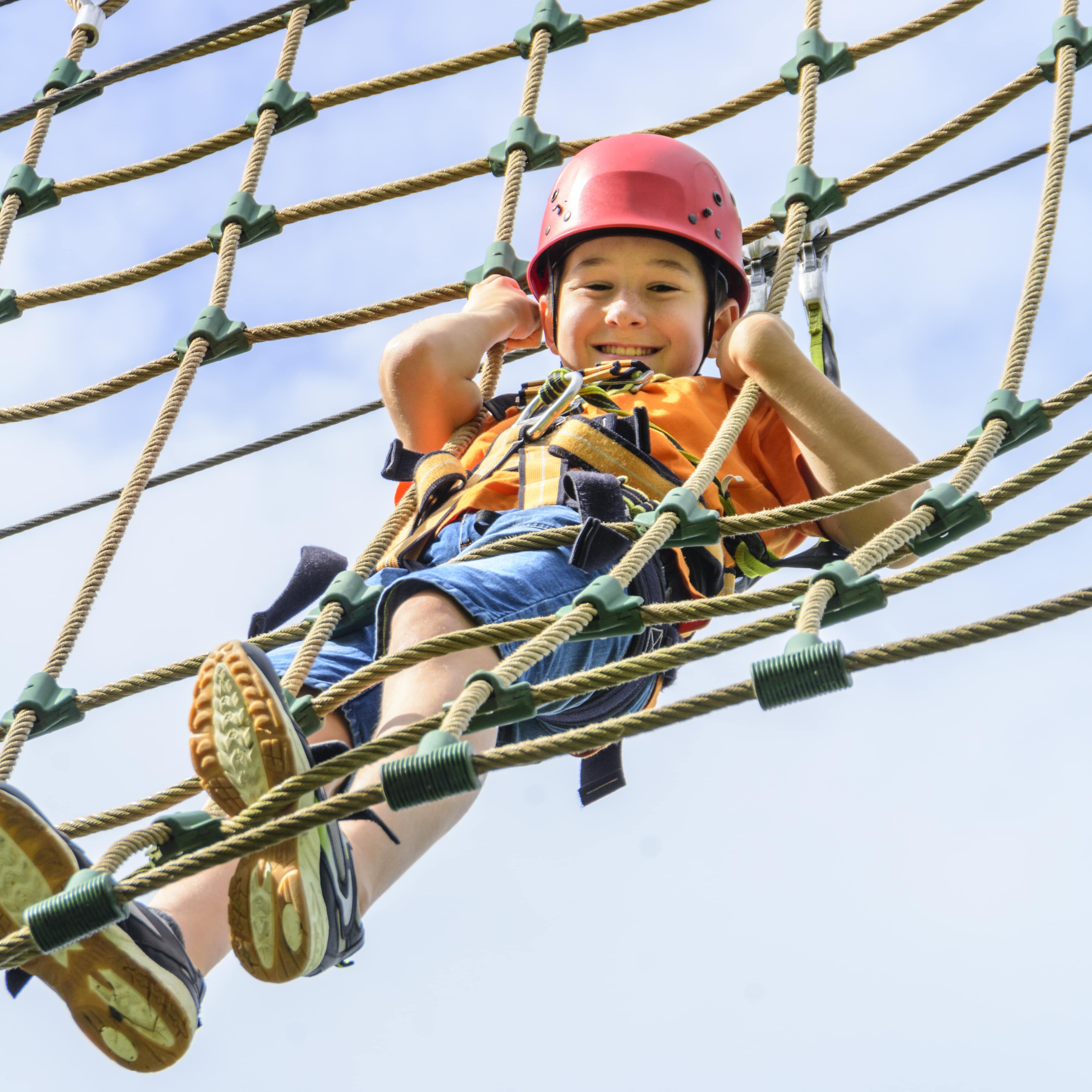 Junge in Sommerkleidung mit Helm und Sicherheitsgurt an einem Kletternetz im Hochseilgarten.