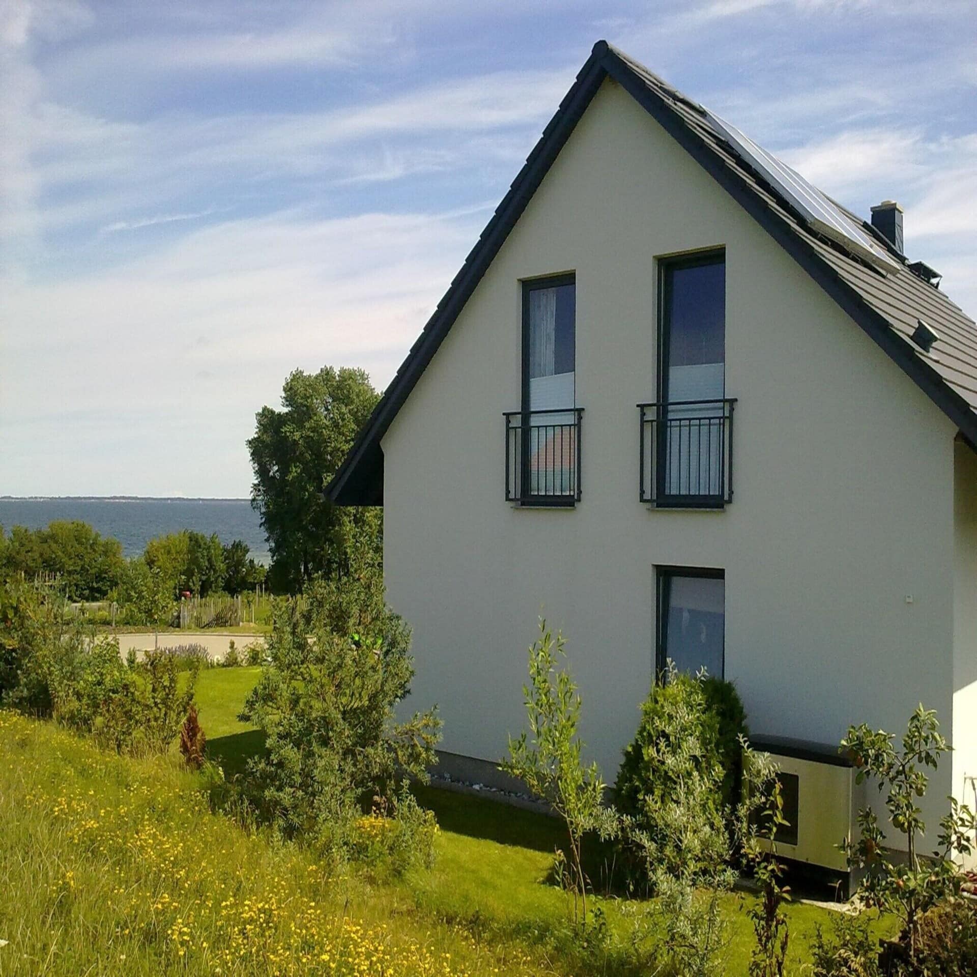 Haus mit Garten, dahinter ist das Meer zu sehen. Die Sonne scheint, es ist Sommer.