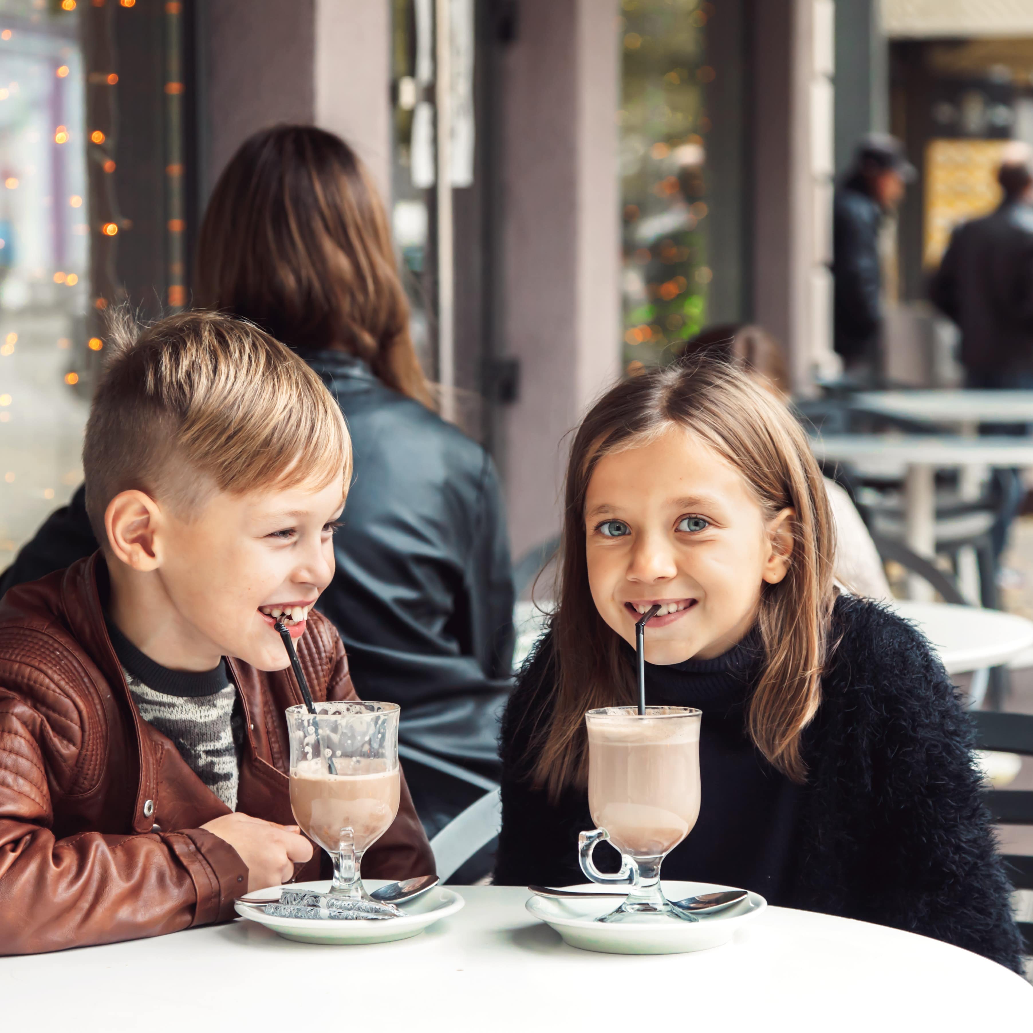   2 Kinder sitzen an einem Tisch in einem Straßencafé und trinken heiße Schokolade mit Strohhalm.