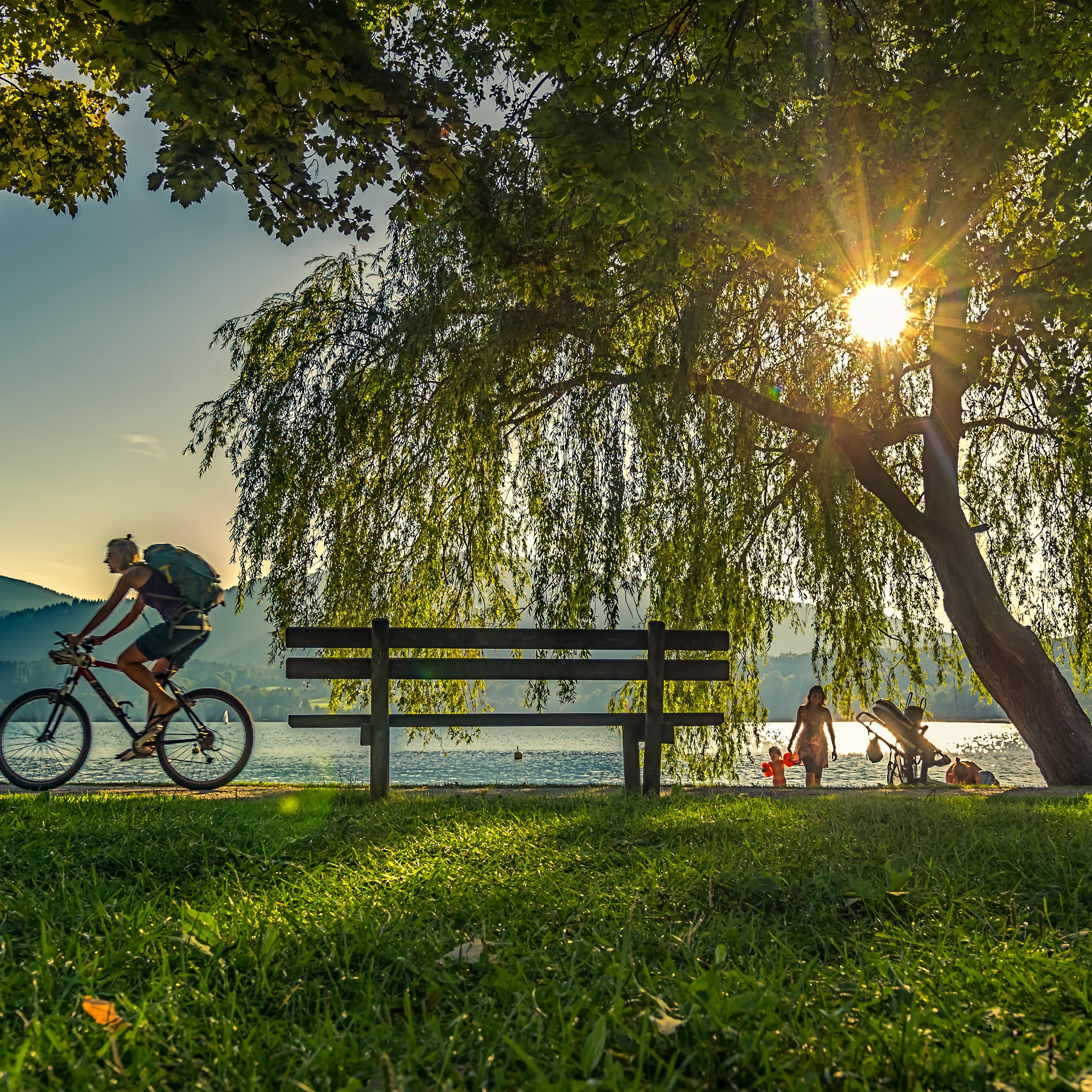 Eine Wiese am See im Sommer. Ein Radfahrer und einige Leute in Badebekleidung am Ufer. 