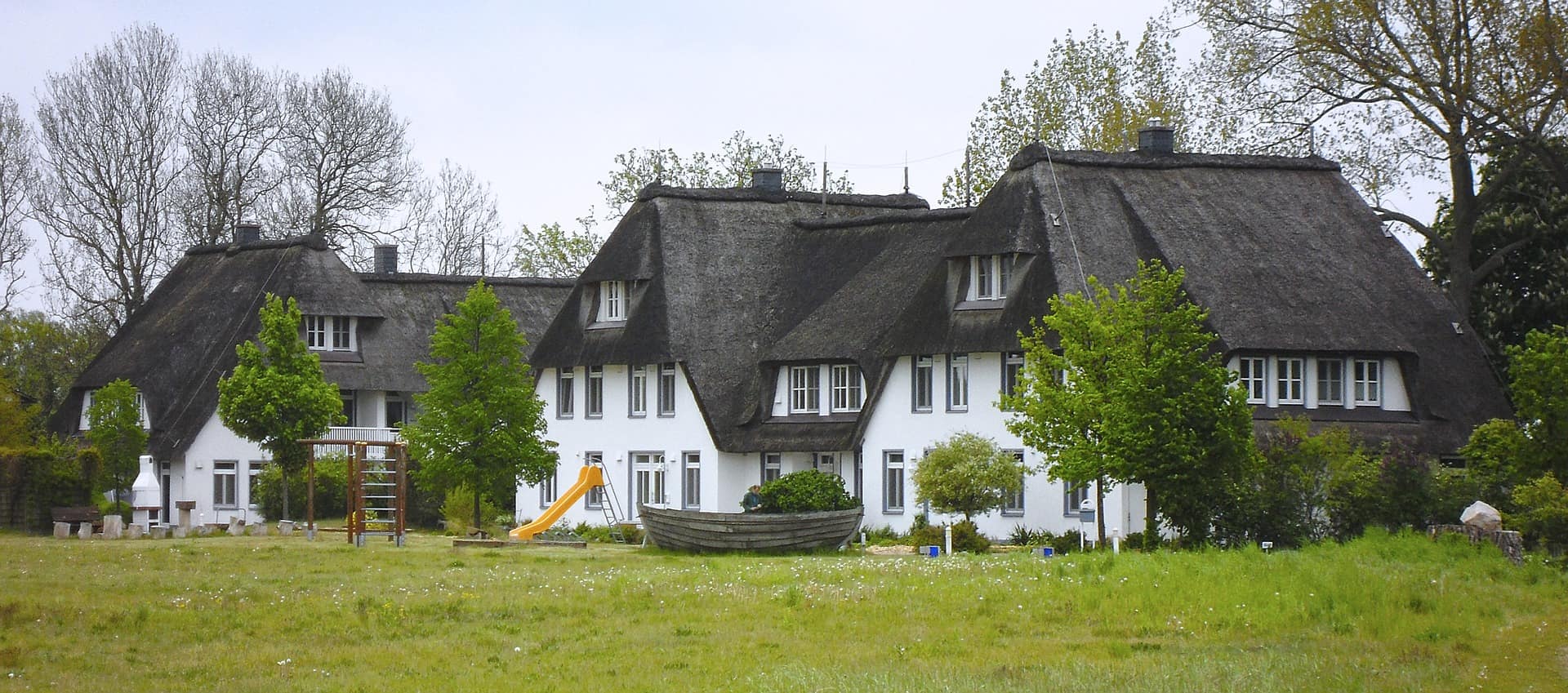 Blick auf große weiße Häuser mit Reetdach und Spielplatz auf eine Wiese