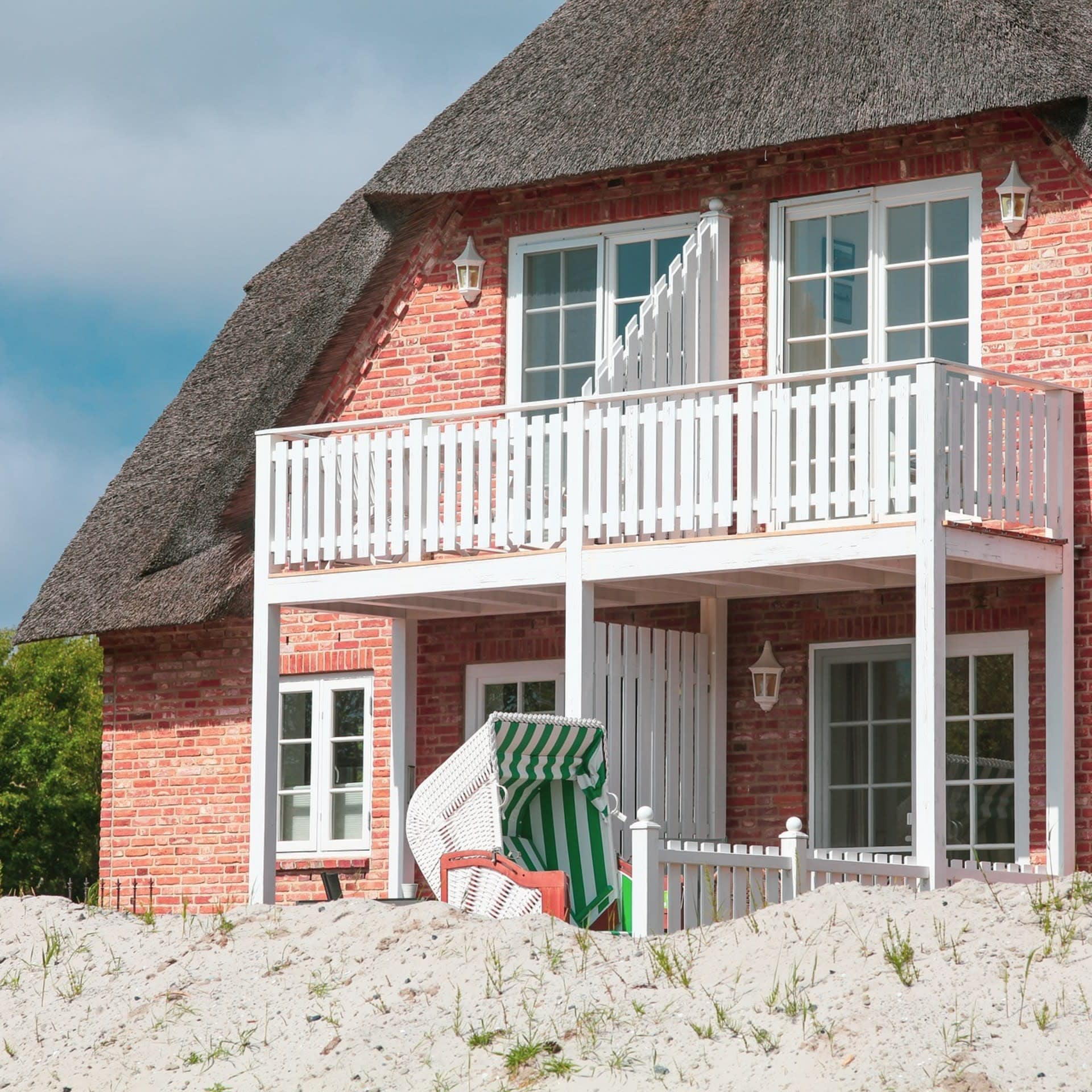 Rotes Friesenhaus mit Reetdach und weißem Balkon. Ein Strandkorb steht im Sand vor dem Haus.