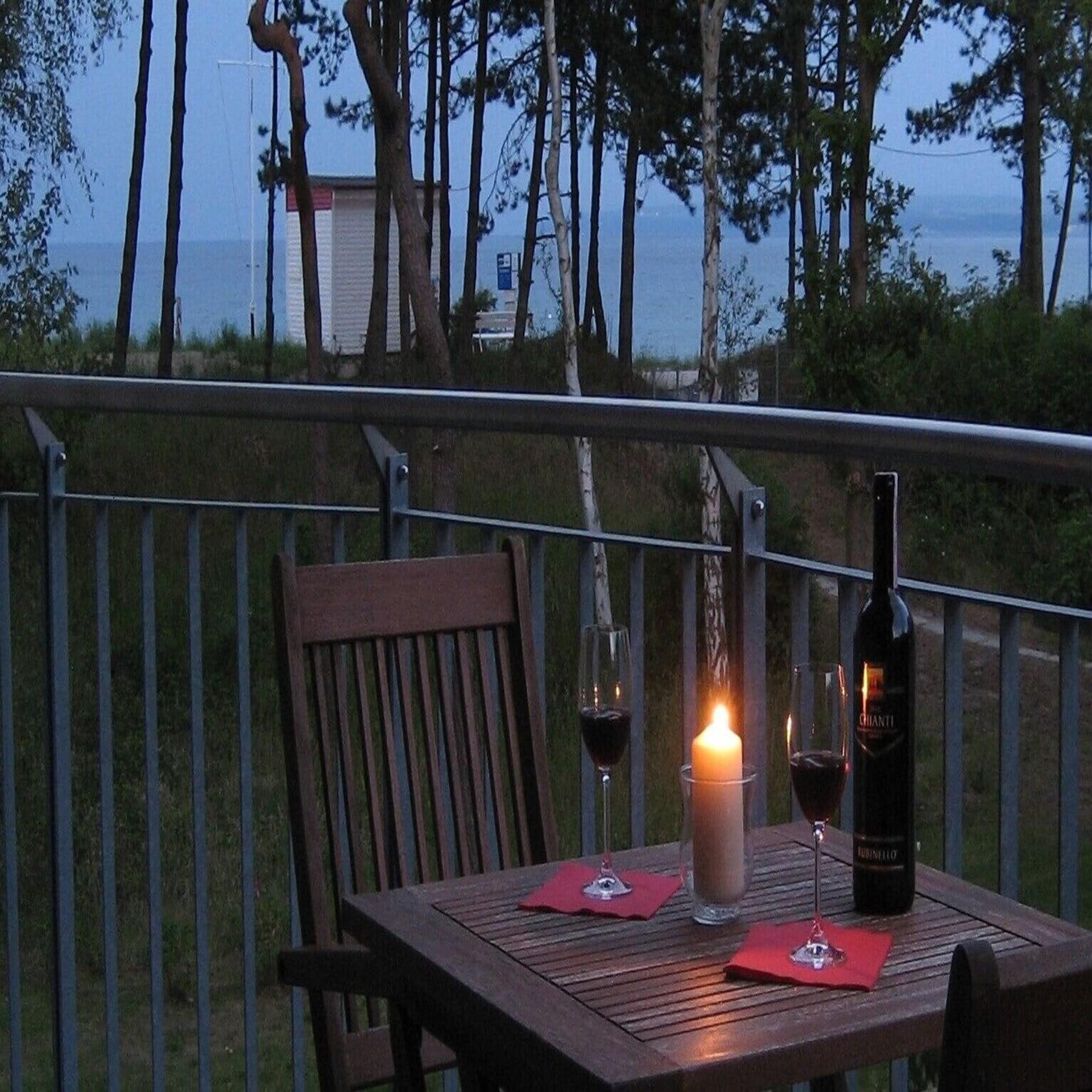 Balkon mit Tisch, Stühlen, Kerze und Wein in der Dämmerung und Blick durch die Bäume auf die Ostsee.