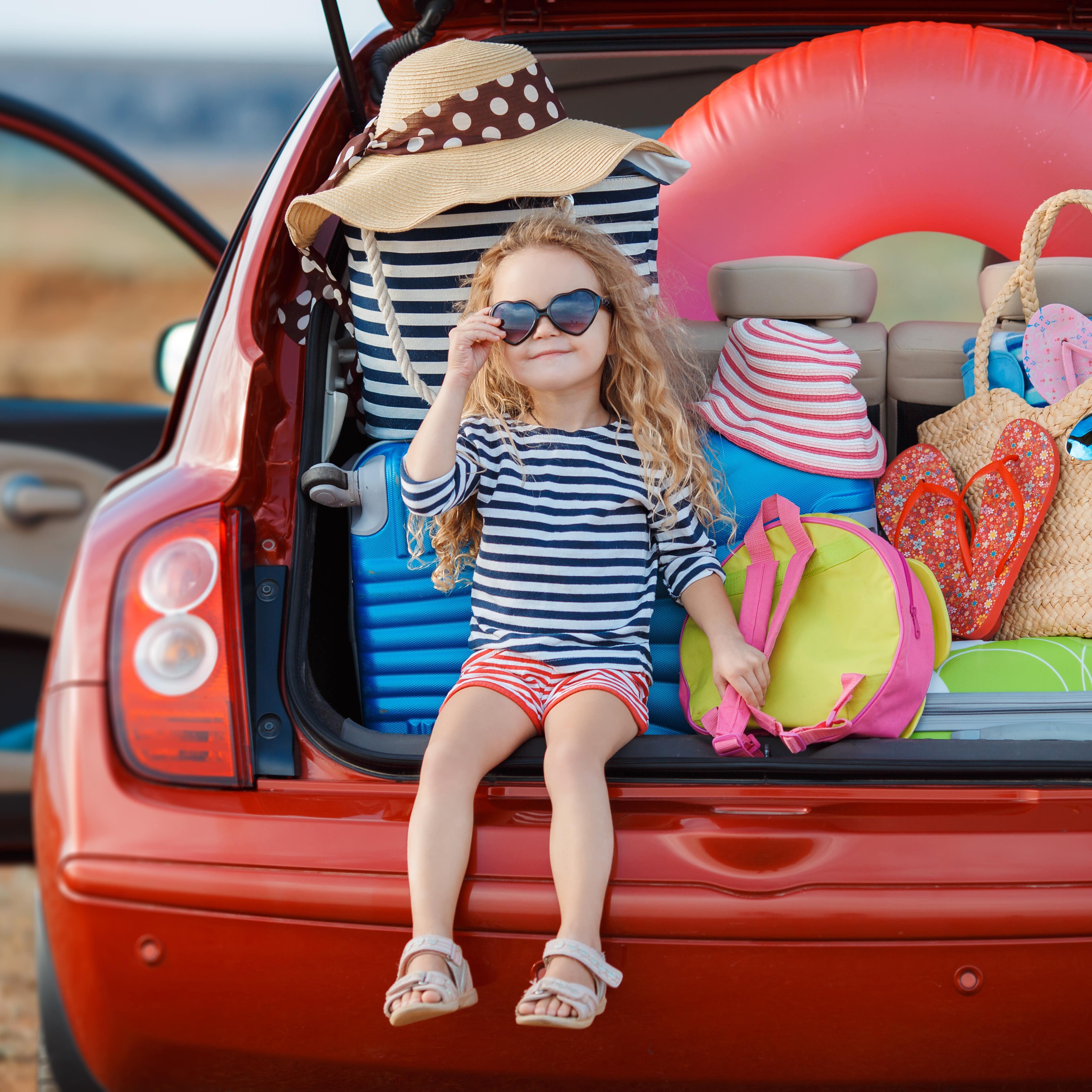 Ein kleines Mädchen in gestreiften Sommersachen sitze in einem vollgepackten Kofferraum eines roten Autos.